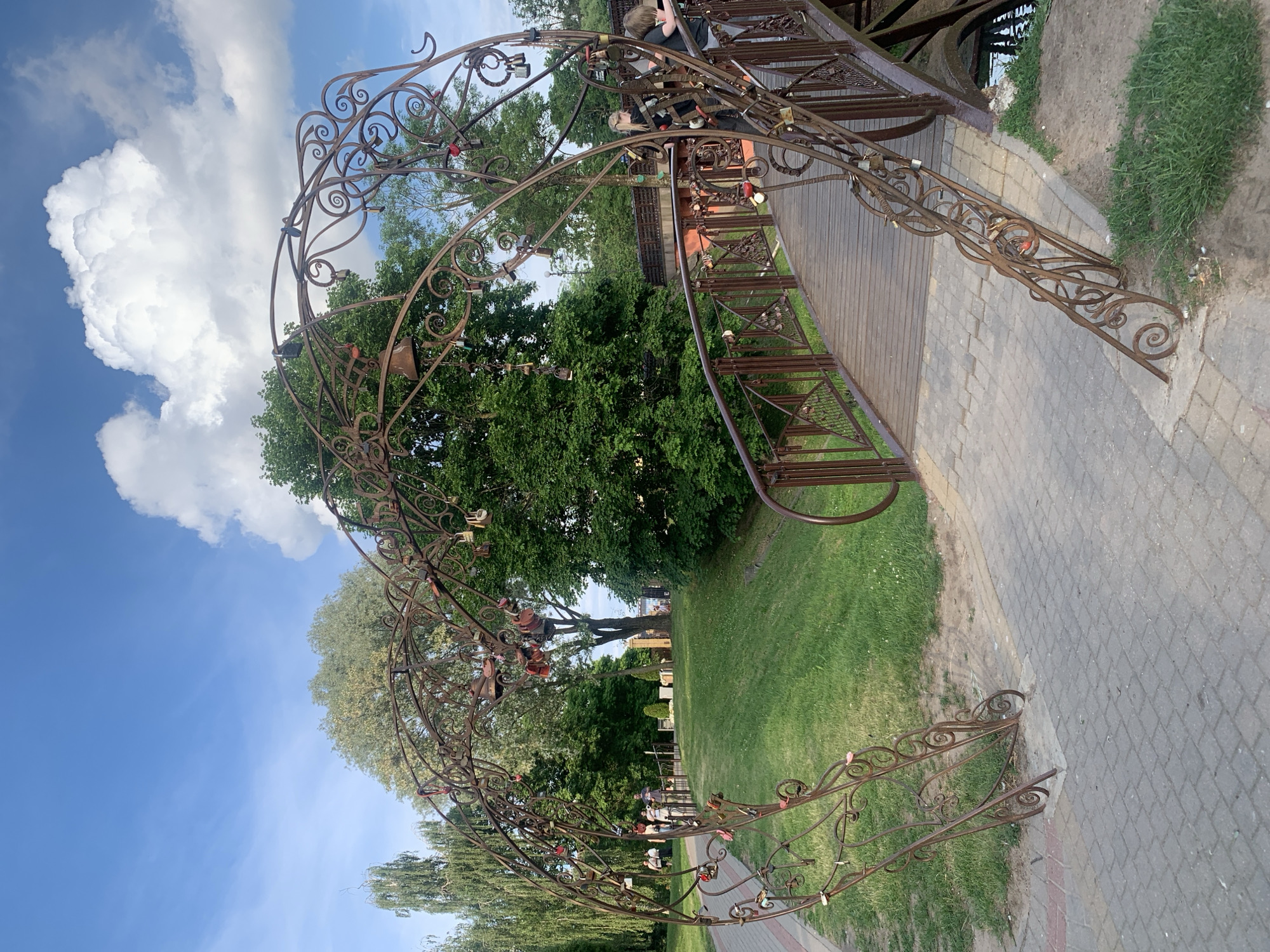 This image depicts a pedestrian park scene in Hrodna, Belarus, centered around a prominent, weathered, dark-brown wrought-iron archway. The arch, styled with numerous scrolls and decorative elements, resembles a large heart or two opposing arches, and is heavily adorned with hundreds of "love locks" and several small bells of various sizes.
The arch spans a light-grey cobblestone pathway that leads to a pedestrian bridge. The bridge features a wooden plank surface and ornate wrought-iron railings matching the style of the archway, also covered in numerous "love locks." A narrow waterway is partially visible beneath the bridge on the right side.
On the bridge, near the right side of the arch, two individuals are visible: a blonde girl in a dark top and a boy with long dark hair, both appearing to be children, leaning against the bridge railing. Further along the cobblestone path to the left of the arch, several other people are present, appearing as indistinct figures strolling in the distance.
The environment consists of well-maintained green grass on gentle slopes, bordered by a variety of mature trees. These include a tall, slender tree with light-colored bark and sparse foliage on the far left, and denser, darker green deciduous trees throughout the background. The sky is bright blue with scattered white clouds, notably a large, prominent cumulus cloud on the upper right side. The scene is illuminated by natural daylight, casting soft shadows, suggesting a clear day, possibly late afternoon.
