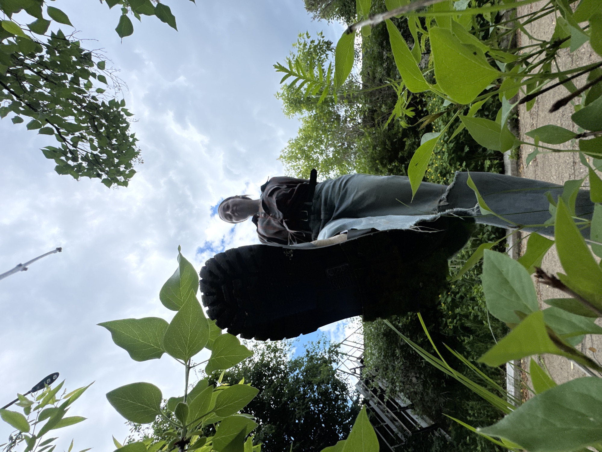 A low-angle shot shows a person standing, with the view from the ground looking up past their boot, legs, and torso. The boot sole fills a significant portion of the lower frame. The person is wearing jeans, and what appears to be a flannel shirt or jacket. The person’s face is visible, though small, appearing against a partially cloudy sky. The surrounding environment consists of lush greenery, including leaves and tree branches framing the shot. A glimpse of a building or structure with metal railings can be seen through the vegetation. It is not possible to determine the specific location as being the city of Chaikovsky, Russia, because there are no elements in the image that would clearly establish the city as the location.