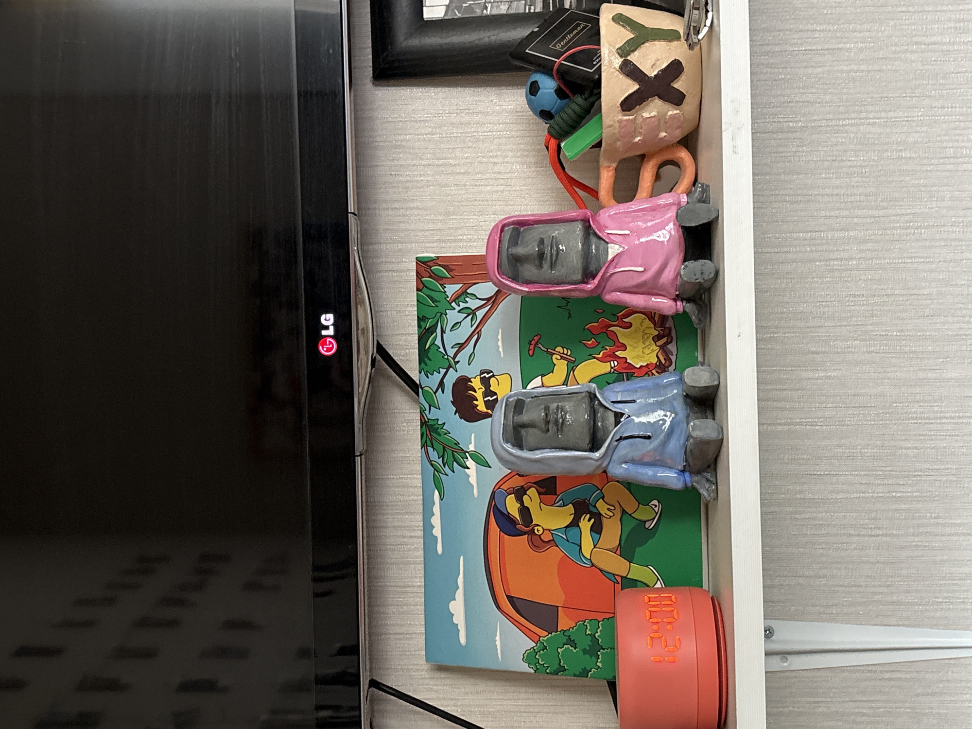 The image shows a shelf with various items. Starting from the left, there is a large, black LG television. To the right of the TV, there is a framed picture and a small container holding various items including a "Gentleman" brand item, a small blue ball, and a green object. Next to these is a stylized mug with "XY" written on it. Below these items, there are two figurines of people wearing pink and blue jackets, respectively. Below the figurines, there is a colorful painting showing cartoon characters in an outdoor setting, complete with a tent and campfire. Finally, at the bottom right of the image, there is a small, orange digital clock displaying "00:28". The location is likely indoors. There is no information to associate this image with the city of Krasnoyarsk, Russia.
