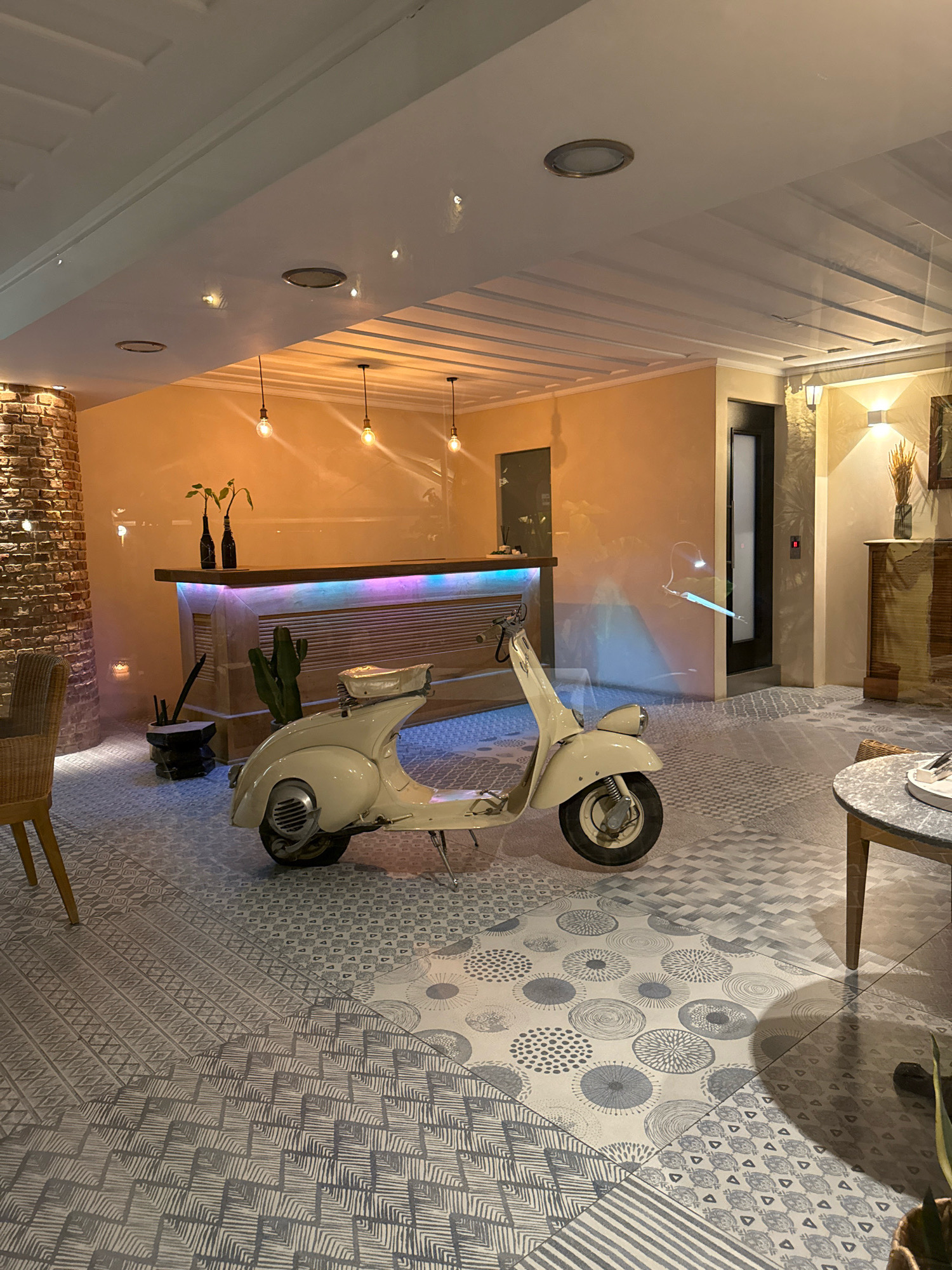 The image depicts an indoor scene, likely a lobby or reception area, with a vintage cream-colored scooter prominently placed on a floor with a complex geometric tile pattern. The scooter is positioned in the foreground, facing left. Behind it, a wooden reception desk is visible, illuminated by colorful LED lights that cast a vibrant glow on its textured surface. The wall behind the desk is painted a warm orange hue. To the left of the reception desk, a section of exposed brick wall adds a rustic element. Several plants are present in pots near the desk and on the floor, including a tall cactus. The ceiling features a modern design with recessed lighting. The overall ambiance suggests a stylish and contemporary establishment with a retro touch. No people are visible in the image.