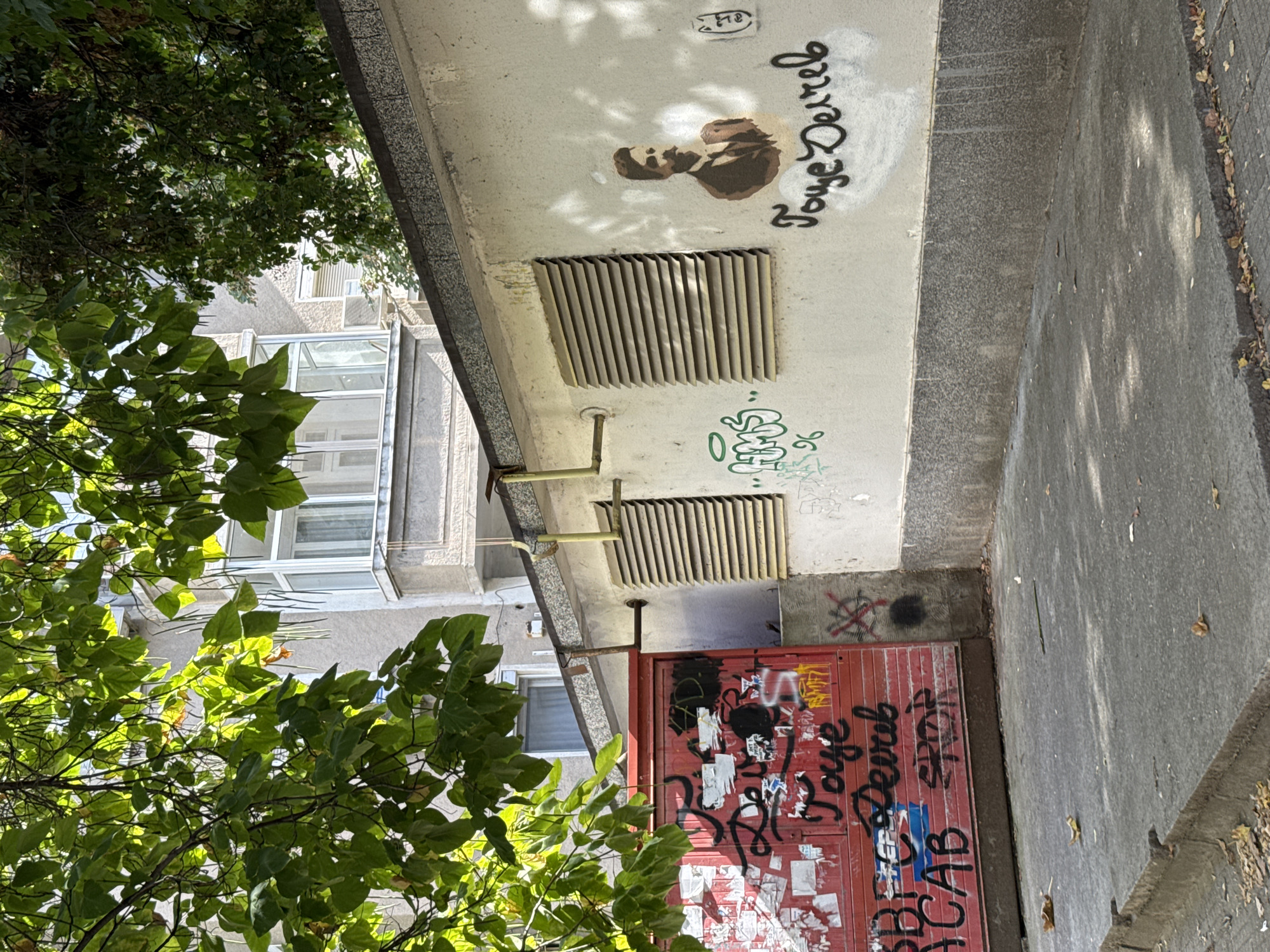 The image depicts a section of a wall with graffiti and a red metal gate in Plovdiv, Bulgaria. The wall is light-colored and appears to be part of a building or an enclosure. On the wall, there is a stencil of a man in a suit and tie, with his name or a phrase written beneath it in black graffiti. To the left of the stencil, there is green graffiti with numbers. To the right, there is another small graffiti tag.
The red metal gate is covered in various tags and stickers, mostly in black and white, with some red and blue elements. Some visible text includes "ACAB" and "SPOR".
The scene is outdoors, with trees and sunlight casting shadows on the wall and the ground. The ground in front of the gate and wall is paved with concrete. The overall impression is an urban environment with signs of vandalism or street art.