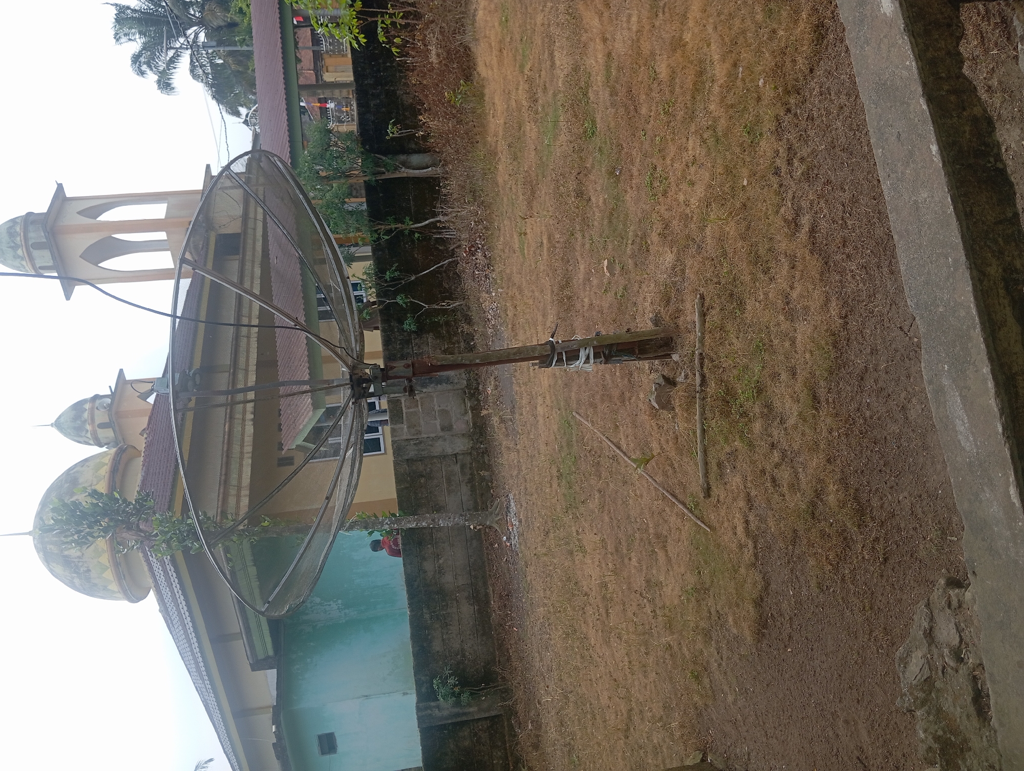 In Rejosari, Indonesia, a satellite dish is positioned in an outdoor area with dry grass and patchy dirt. A concrete wall runs horizontally in the background, behind which a building with a light yellow facade and a domed roof is visible, likely a mosque, featuring architectural elements of Islamic design. To the left of the building, a section of a lighter blue wall is present. A lone individual wearing a red shirt is seen standing near the concrete wall, facing away from the camera. The satellite dish itself is a large, dark mesh parabola mounted on a metal pole, secured with white tape. The environment appears somewhat unkempt, with scattered dry leaves and debris on the ground.