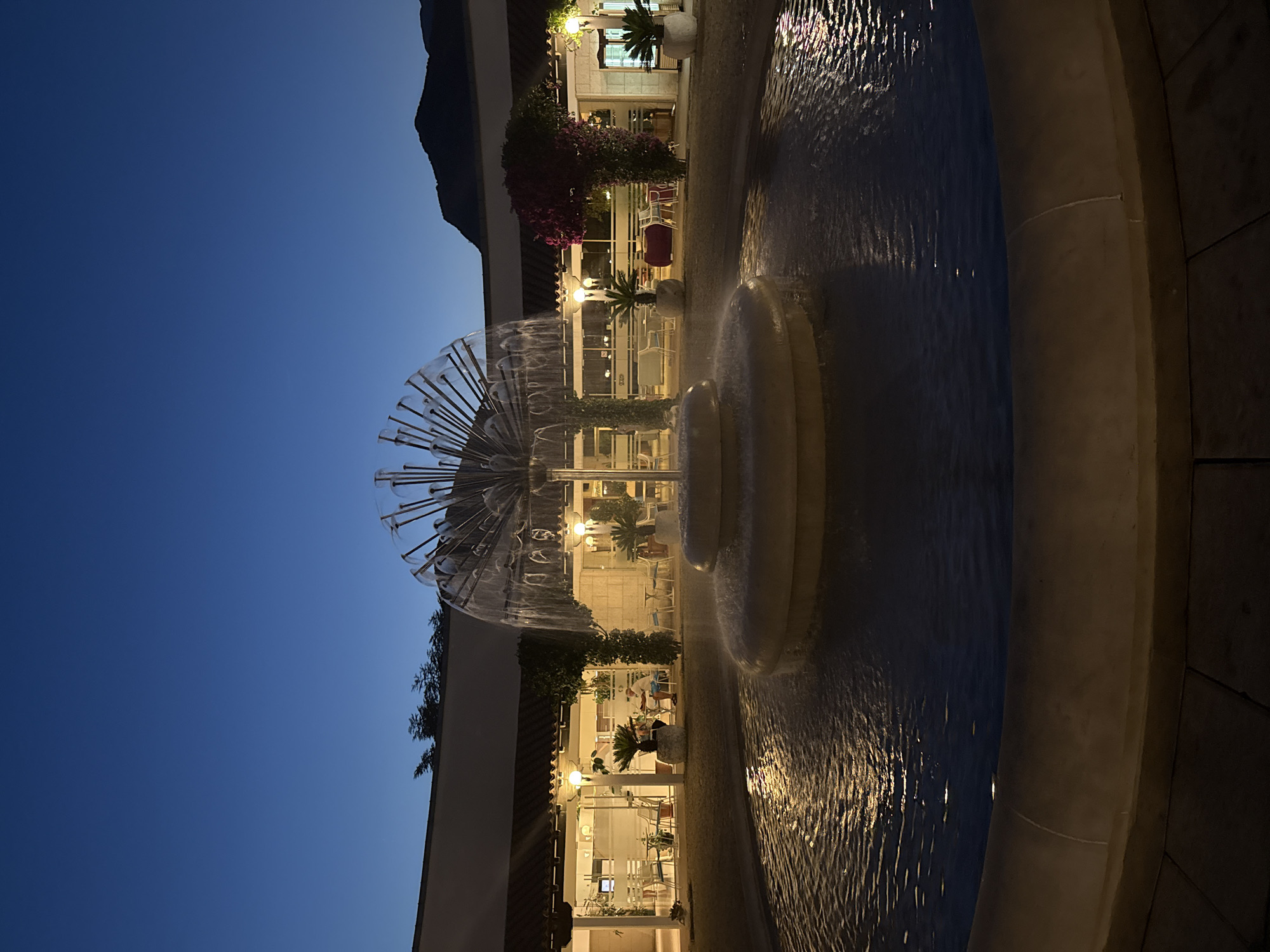 The image shows a circular outdoor fountain with a modern, sculptural water feature in the foreground. The fountain is illuminated by warm light reflecting off the rippling water. In the background, there is a covered walkway with outdoor seating and tables, suggesting a cafe or restaurant area. Lush green plants and a vibrant pink flowering bush are visible along the walkway. Several people are seated at tables, and the scene is bathed in the dim light of twilight, with a dark blue sky above and silhouetted mountains in the far distance. The location is identified as Budva, Montenegro.