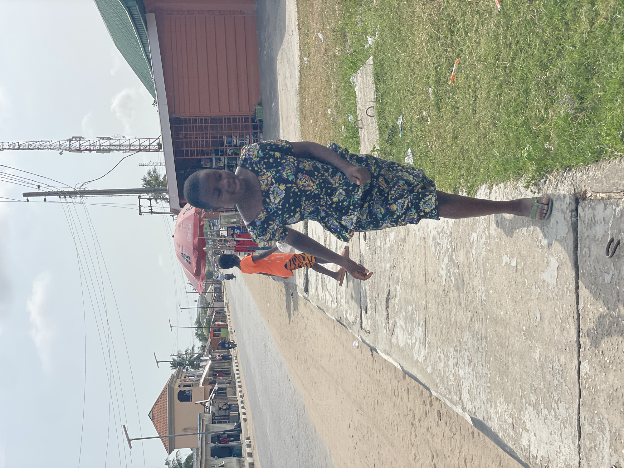 The image shows a woman in a floral dress standing on a concrete ledge next to a sandy road in Badagry, Nigeria. A boy in an orange shirt and tiger-striped shorts walks on the road to the left of the woman. A red Coca-Cola umbrella is visible behind the boy. In the background, there are buildings, utility poles, and a communication tower. The environment appears to be urban with commercial buildings and vegetation nearby. There is some litter on the ground.