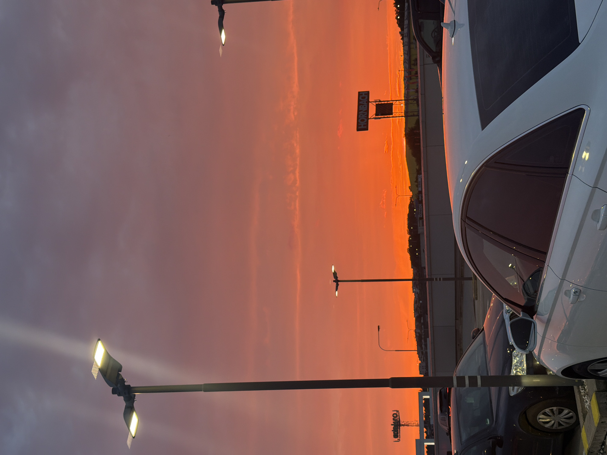 The image depicts an outdoor scene at dusk, likely a parking lot or commercial area. A vibrant orange and pink sunset dominates the sky. In the foreground, a white sedan is parked, with its rear visible. To the left, a dark-colored sedan is also parked. Several streetlights are present throughout the scene, with one in the foreground on the left casting a bright light upwards and another in the mid-ground on the right. In the background, a sign that reads "HORNBACH" is visible, indicating a commercial establishment. Other buildings and structures are also present in the distance. The overall impression is that of an urban landscape at sunset. The specific location in the city of Prague, Czechia cannot be determined from the visual information alone.