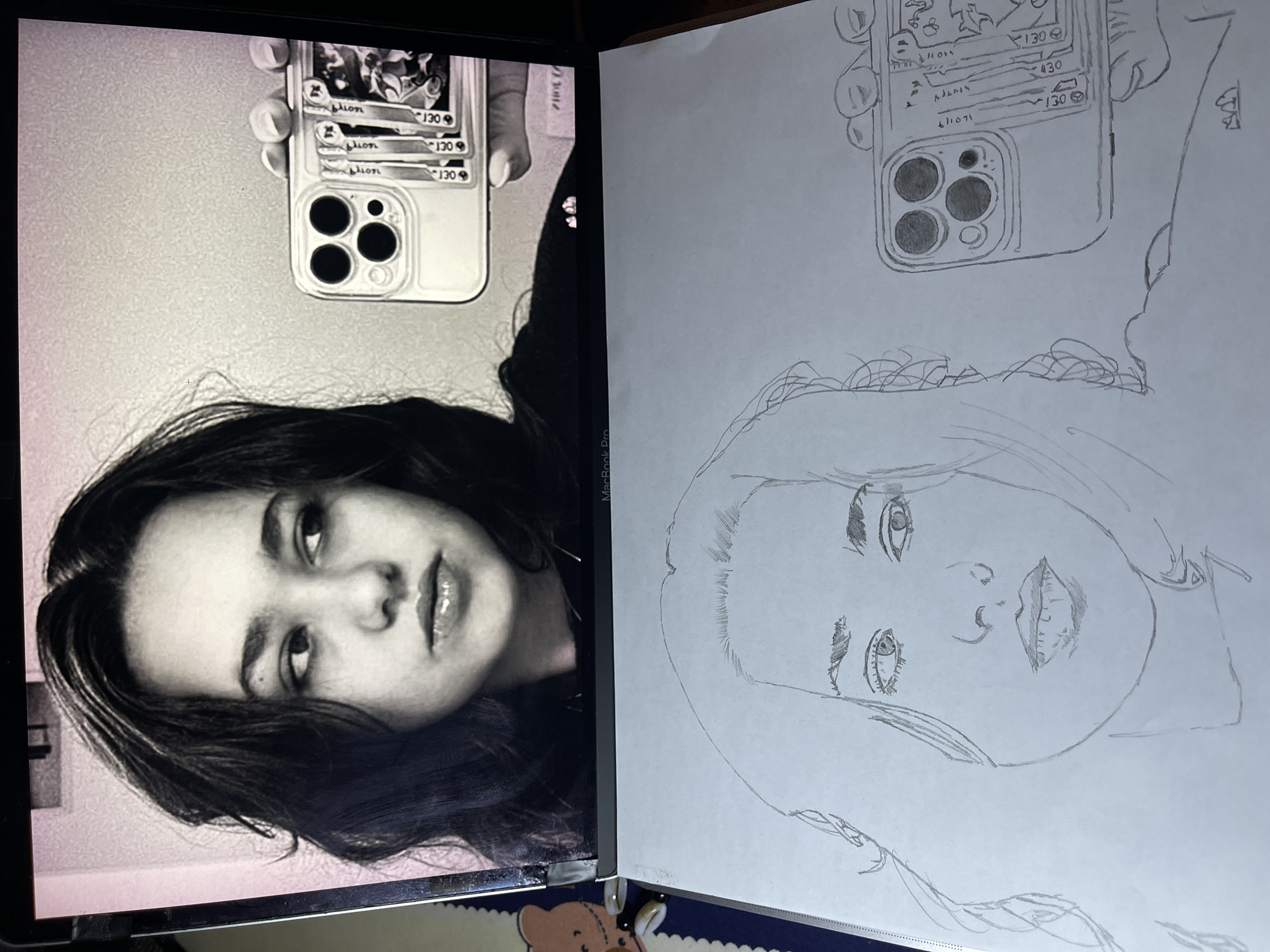 The image shows a side-by-side comparison of a photograph and a pencil sketch. The photograph, displayed on a laptop screen, depicts a woman taking a selfie. She holds a smartphone with a distinctive case featuring what appear to be trading cards attached to the back. Her hair is dark and loose, framing her face. The sketch replicates the same subject and composition, capturing the woman's likeness, the smartphone, and her pose. The setting is indoors with some details visible in the foreground of the physical scene. The city of Jos, Nigeria, is not visibly depicted in the image, so its location is unknown.