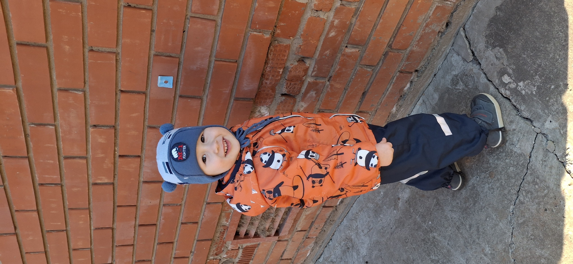 A young child stands beside a brick wall and concrete pavement. The wall is made of red bricks with visible mortar. A small blue square is attached to the wall. The pavement is gray concrete with visible cracks and signs of wear.
The child is wearing a blue and gray hat with ear flaps, a jacket with an orange background and panda bear pattern, and blue pants with white reflective stripes. They also wear gray sneakers with laces. The child is smiling.
The location is likely in the city of Chaikovsky, Russia, based on geographical data and metadata associated with the image.
