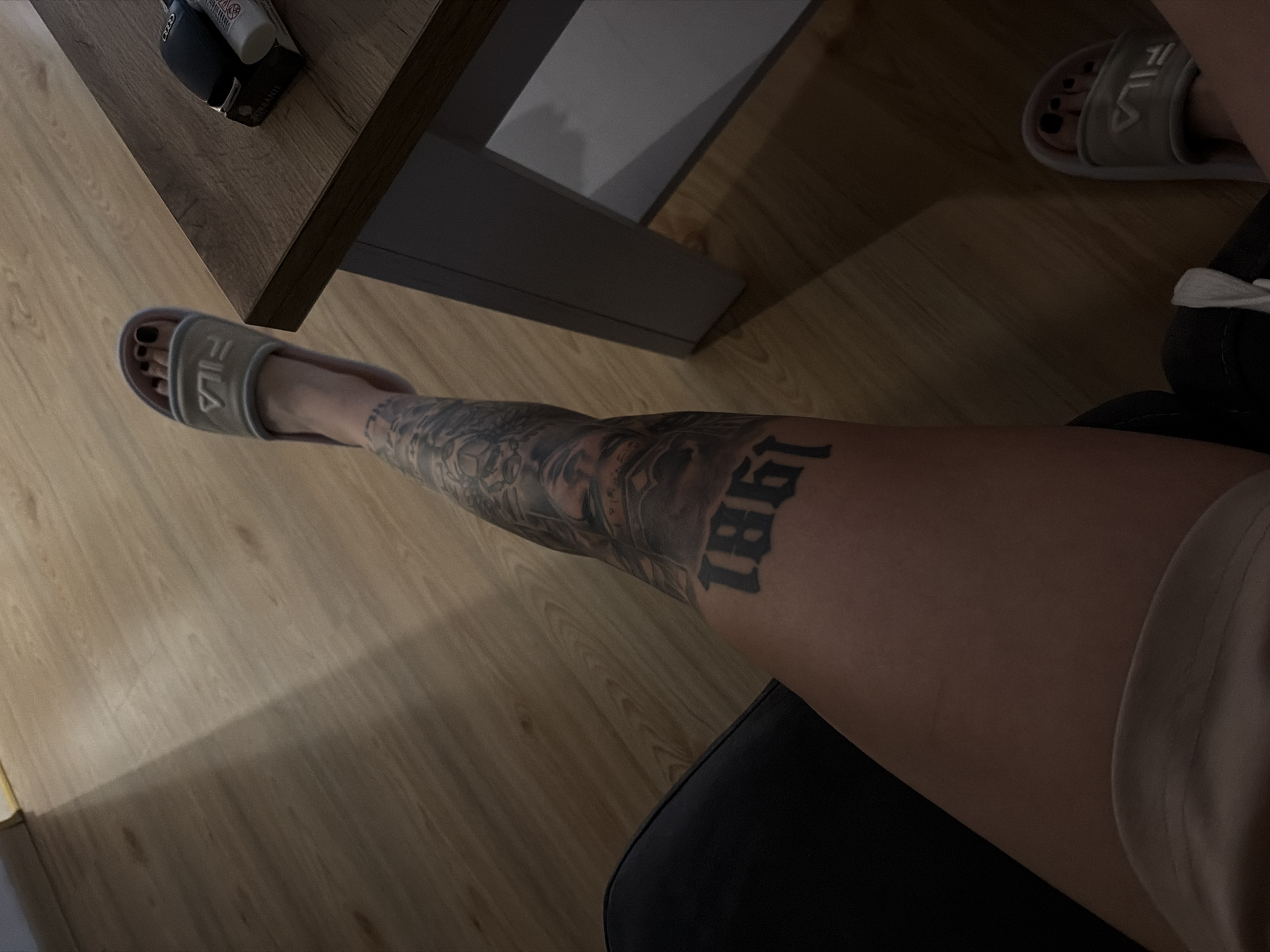 The image shows a person's legs and feet, wearing Fila slide sandals. The person has tattoos on their leg, including the number "1861". The scene is indoors, with a wooden floor and a glimpse of a table with some items on it. There is no indication of the location being Plovdiv, Bulgaria.
