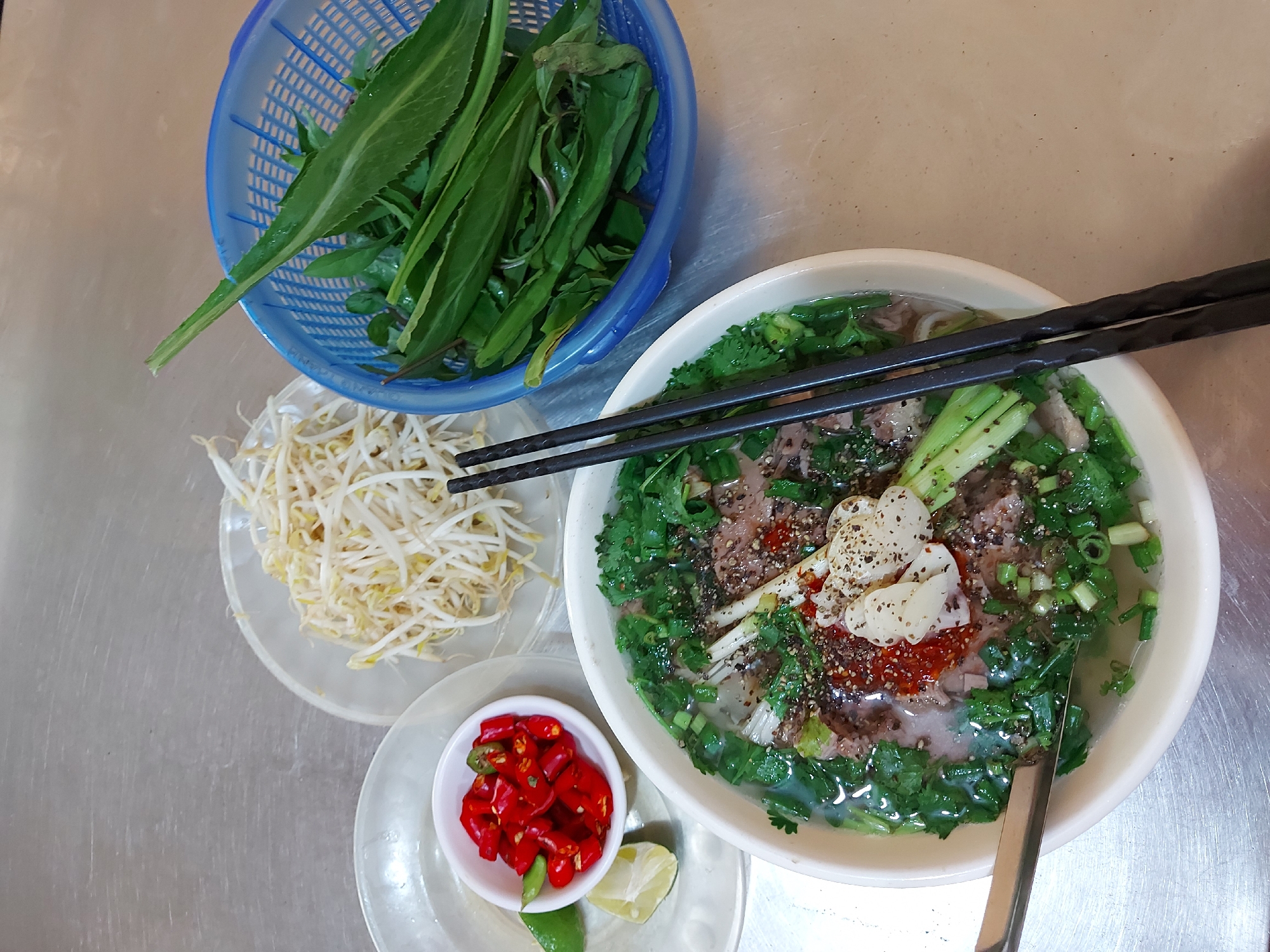An overhead shot shows a bowl of Vietnamese pho, a popular noodle soup, and its accompaniments, arranged on a stainless steel surface. The pho, served in a white ceramic bowl, contains a rich broth, thin slices of beef, noodles, chopped green onions, and fresh cilantro. A dollop of chili paste and some sliced garlic cloves are visible on top of the soup. Two black chopsticks rest across the bowl. To the left of the pho, a small white bowl holds a generous portion of chopped red chili peppers and two lime wedges. Behind the pho, a clear plastic plate displays a mound of fresh bean sprouts. To the right, a blue plastic basket is filled with a variety of fresh green herbs, likely for adding to the pho. The stainless steel surface suggests a food stall or restaurant setting, common in cities like Da Nang, Vietnam. The lighting is natural, likely from an overhead source, casting minimal shadows. The composition is flat and direct, with no people or visible action.