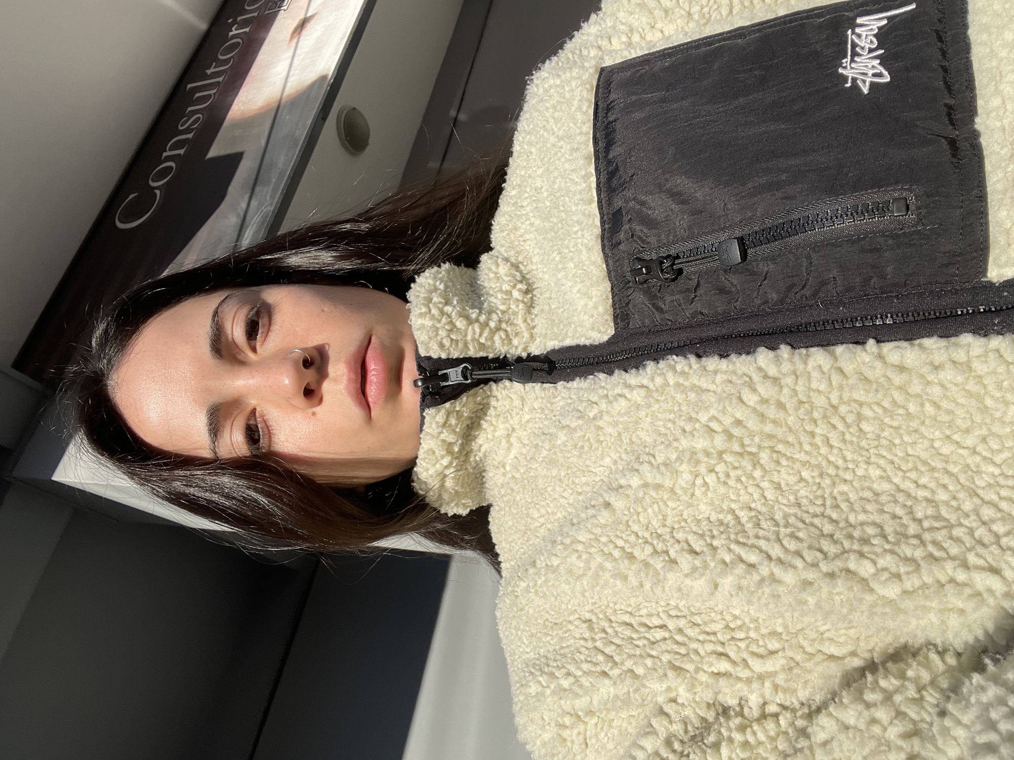 The image shows a woman wearing a light-colored, fleece jacket with a black pocket bearing the "Stussy" logo. The woman has dark hair and fair skin. The image is taken indoors, with a sign reading "Consultorio" visible in the background. Given the location query, this would likely be a doctor's or medical office in Ushuaia, Argentina. The lighting suggests it's daytime. The image is rotated, with the woman appearing sideways.