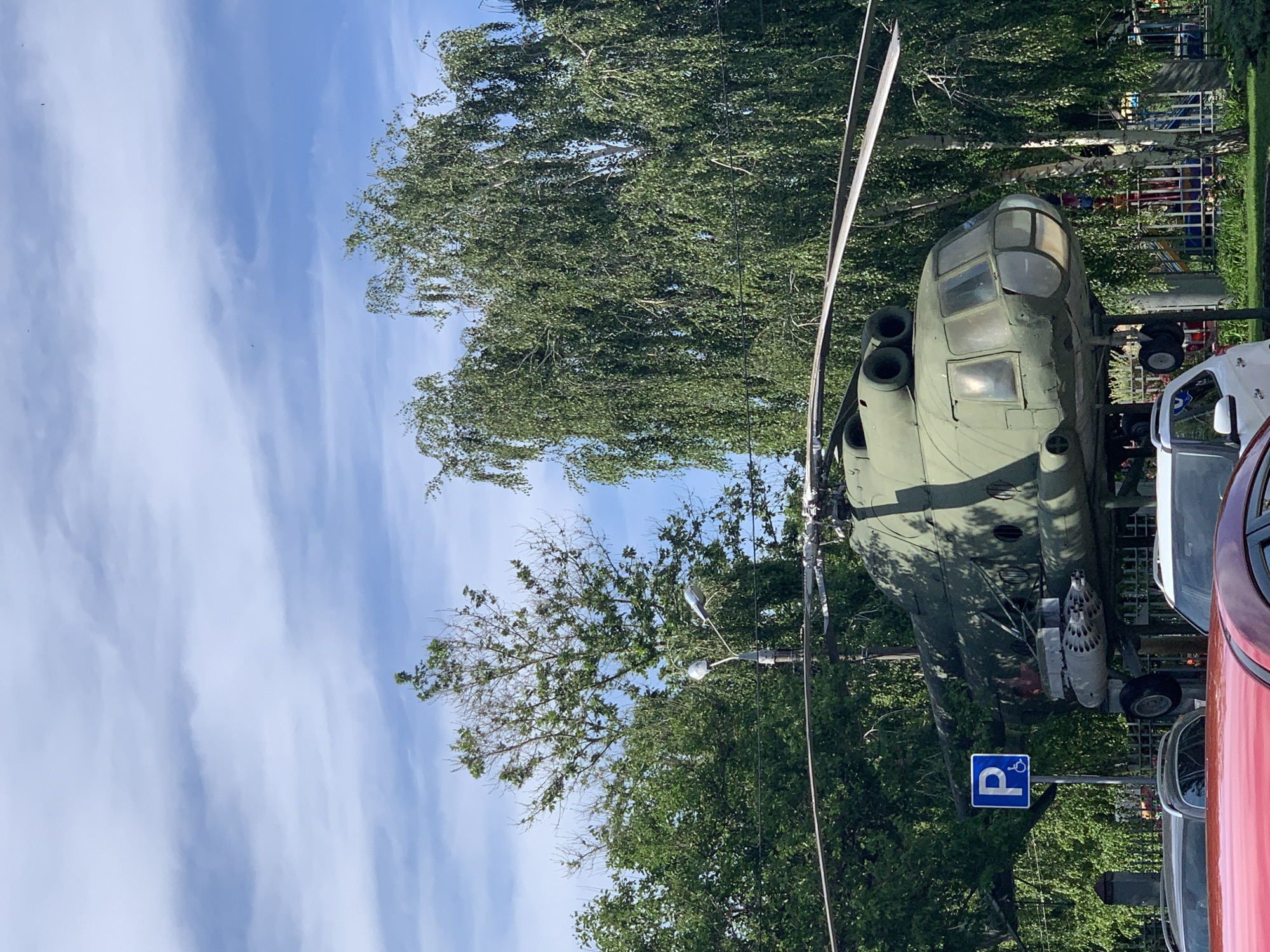 A clear, bright daytime image shows a large, olive-green military-style helicopter displayed outdoors, likely as a monument or exhibit. The helicopter, possibly a transport variant given its size and cockpit design, features a prominent main rotor blade extending upwards and two large engine exhaust ports visible on its top. A pod with multiple missile or rocket tubes is mounted on its side, suggesting its combat capability.
The scene is set under a wide expanse of bright blue sky with wispy white clouds. Lush green deciduous trees, some with light-colored bark typical of birch trees, surround the helicopter, creating a natural backdrop. Sunlight casts subtle shadows, indicating a sunny day, likely late morning or early afternoon.
To the right of the helicopter, parts of two cars are visible: a white vehicle and a red one, suggesting the display is situated near a road or parking area. A blue square sign with a white capital 'P' and a white wheelchair symbol below it stands in front of the helicopter, indicating an accessible parking spot nearby. A street lamp post is also partially visible among the trees. No people are present in the image. The overall atmosphere is calm and serene.