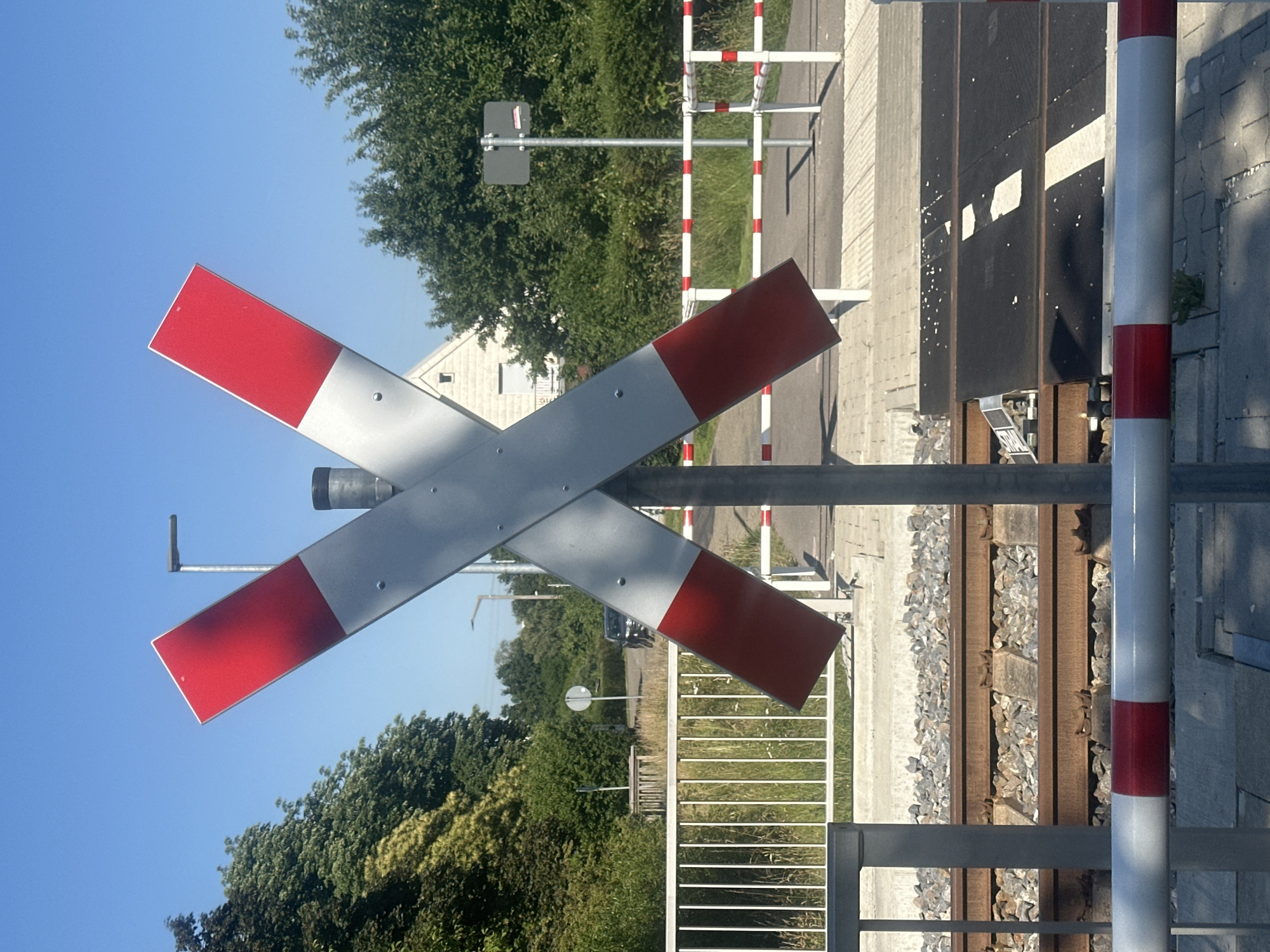 The image shows a railroad crossing in an outdoor environment, likely during daylight with clear skies. The primary subject is a large, red and white striped X-shaped sign, commonly used to indicate a railroad crossing. This sign is mounted on a vertical metal pole. In the background, there are train tracks with ballast stones. Beyond the tracks, a white metal fence with red and white striped horizontal bars stretches across the scene, suggesting a barrier for pedestrian or vehicle traffic. There are green trees and bushes on either side of the railroad crossing. A residential building with a light-colored facade and a pitched roof is visible among the trees on the right side of the image. A lamppost is also present in the background. The ground consists of paved surfaces and gravel. No people or vehicles are prominently featured in the image.