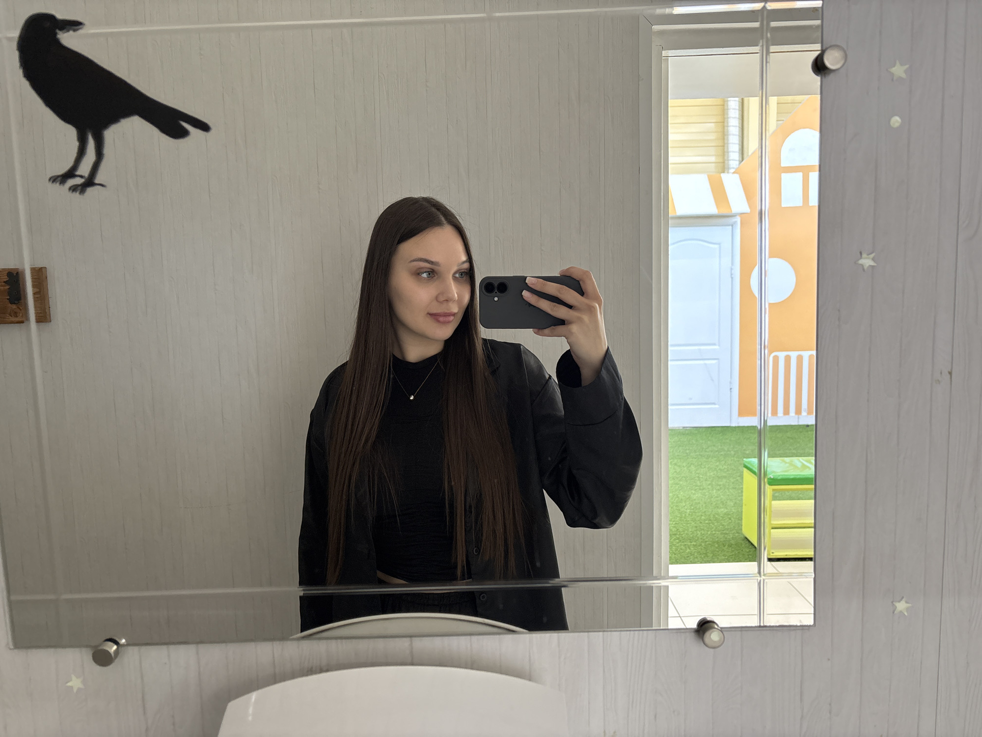 A woman is taking a selfie in a bathroom mirror. She has long dark brown hair and is wearing a black top and a black jacket. In the mirror, a crow sticker is visible on the left side, and a glimpse of what appears to be a children's play area with artificial grass, a bench and a playhouse is visible through an open door or window on the right. A sink is visible at the bottom of the mirror. The setting is Chaikovsky, Russia, as implied by geotag information.