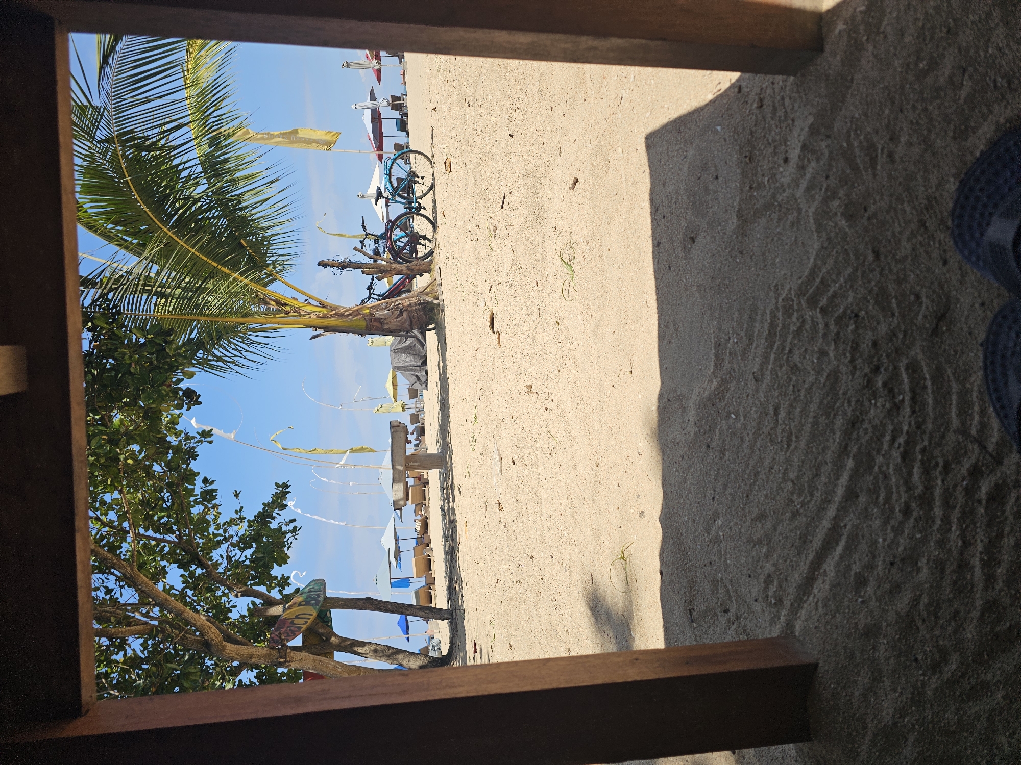 The image captures a serene beach scene in Denpasar, Indonesia, viewed from beneath a wooden structure. In the foreground, coarse sand covers the ground, with tire tracks imprinted from a vehicle. Two dark-colored flip-flops are visible at the very bottom of the frame.
The mid-ground reveals a stretch of sandy beach dotted with several beach umbrellas and lounge chairs, some of which are unoccupied. A wooden bench or table is positioned centrally on the sand. Two bicycles, one red and one blue, are parked near a palm tree. A sign with colorful lettering, partially obscured, appears to indicate "Beach Bar."
The background consists of a clear blue sky and the distant ocean, though the water itself is not prominently visible. The overall atmosphere is peaceful and inviting, suggesting a typical day at a tourist-friendly beach. The presence of the wooden structure in the foreground implies the viewpoint is from a beach hut, cafe, or similar establishment.