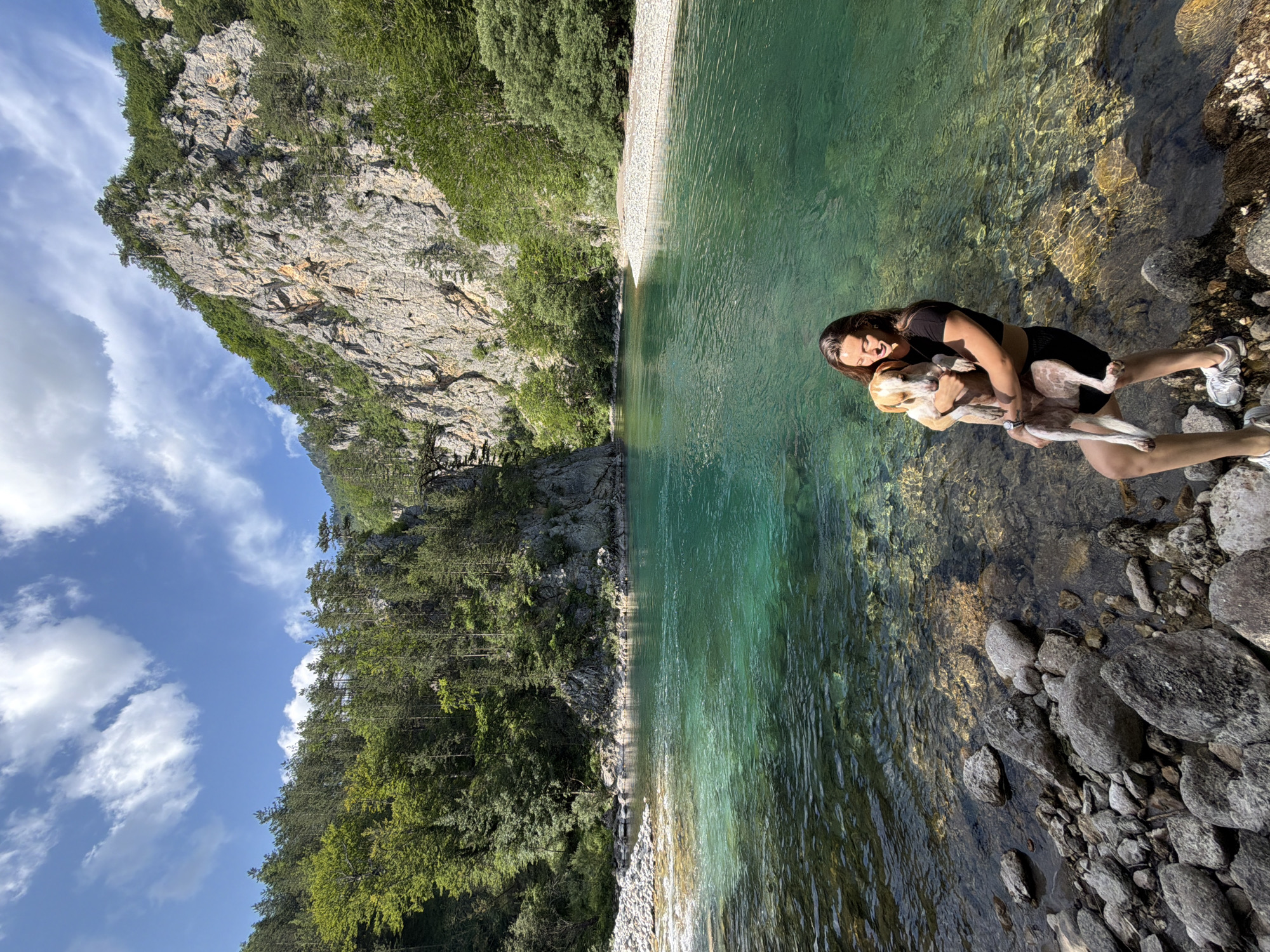 A young woman with brown hair, wearing a black crop top and black shorts, is holding a light brown and white dog in her arms by the edge of a vibrant turquoise river. The river has a rocky bed visible through the clear water and is surrounded by lush green trees and tall, rugged cliffs. The sky above is blue with scattered white clouds. The location is identified as the city of Bistrica, Montenegro.