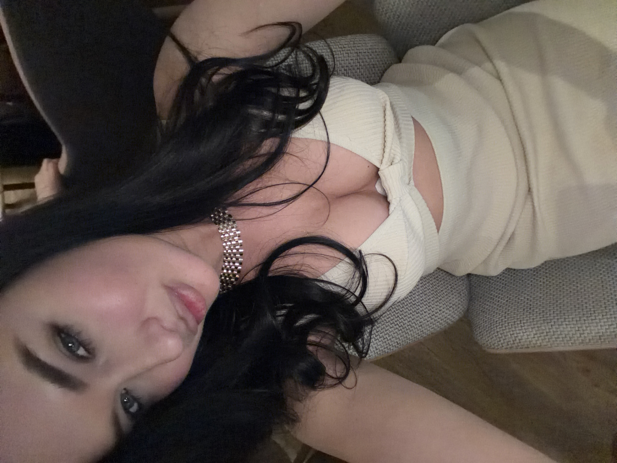 A woman with long, dark hair is reclining in a gray upholstered chair. She is wearing a light-colored, possibly cream or beige, dress with a low neckline, and a silver necklace. She is positioned at an angle with her head near the bottom left corner of the frame, looking towards the camera. The background consists of a dark area on the left and the arm of the chair on the right, along with a glimpse of a wood floor. There are no features of this image to indicate that it was taken in Moscow, Russia.