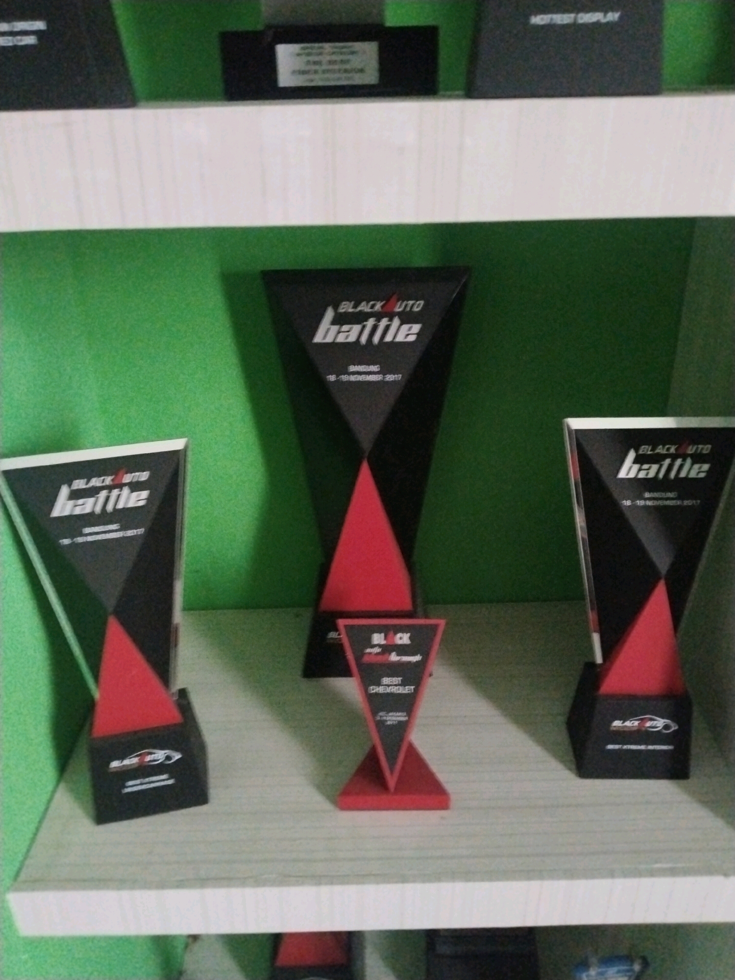 On a white shelf against a green wall in a room, three trophies are displayed. The trophy on the left is black and red, with "BLACK AUTO battle" printed on it. The trophy in the center is also black and red and appears to be a smaller version of the one on the left, with "BLACK AUTO battle" also visible. To the right, another black and red trophy is shown, with "BLACK AUTO battle" printed on it. The room appears to be an indoor setting, possibly a display area or a shelf in a home or office.