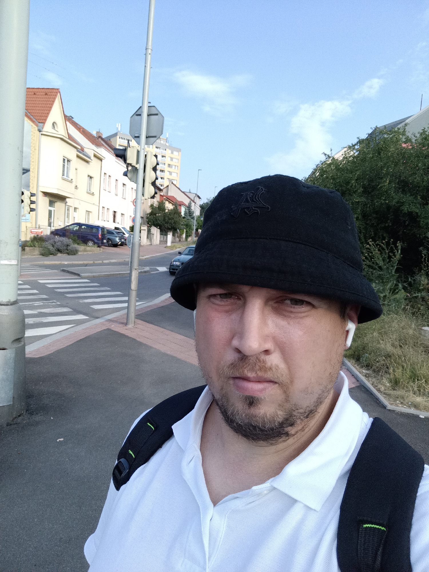The media file is a selfie taken outdoors in a city setting. The main subject is a man wearing a black bucket hat, a white polo shirt, and a black backpack. He has a light beard and is wearing white wireless earbuds. The background shows a street with a pedestrian crossing, traffic lights, and several buildings, some of which appear to be residential. There are cars visible on the street.
The scene appears to be a city street in Prague, Czechia, during the daytime. The sky is blue with some light clouds, suggesting fair weather. The lighting indicates it is likely late afternoon or early evening. The man's expression is neutral. There is no visible text in the image.