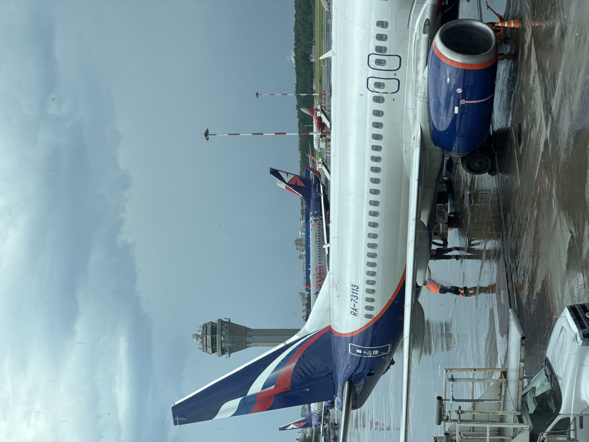 This is a photograph taken at an airport, likely Pulkovo Airport in Saint Petersburg, Russia, on a cloudy day. In the foreground, the tail and fuselage of a white passenger jet with blue and red accents are visible. The registration number RA-73113 is clearly marked on the fuselage. Two ground crew members, one wearing an orange high-visibility vest and dark pants, are working near the aircraft's engine. The tarmac is wet, suggesting recent rain. In the background, several other aircraft are parked, and a control tower with a distinctive honeycomb-like structure stands tall against the sky. Red and white striped light poles are also present. The overall atmosphere is subdued due to the overcast weather.