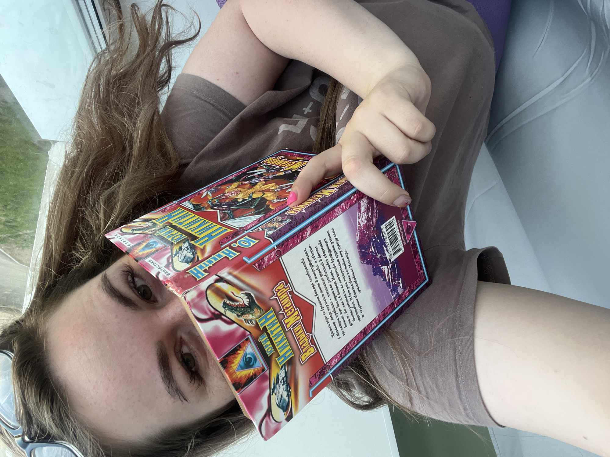 A person is holding a book in front of their face. The book, titled "Владыки Мегамира" ("Masters of Megaworld") by Yuri Nikitin, features a cover with fantasy illustrations. The person, likely female, has brown hair and is wearing a brown T-shirt. Part of their face, including their eyes, is visible behind the book. A window is visible behind them. There is no indication in the image that would specify the location as Chaikovsky, Russia.