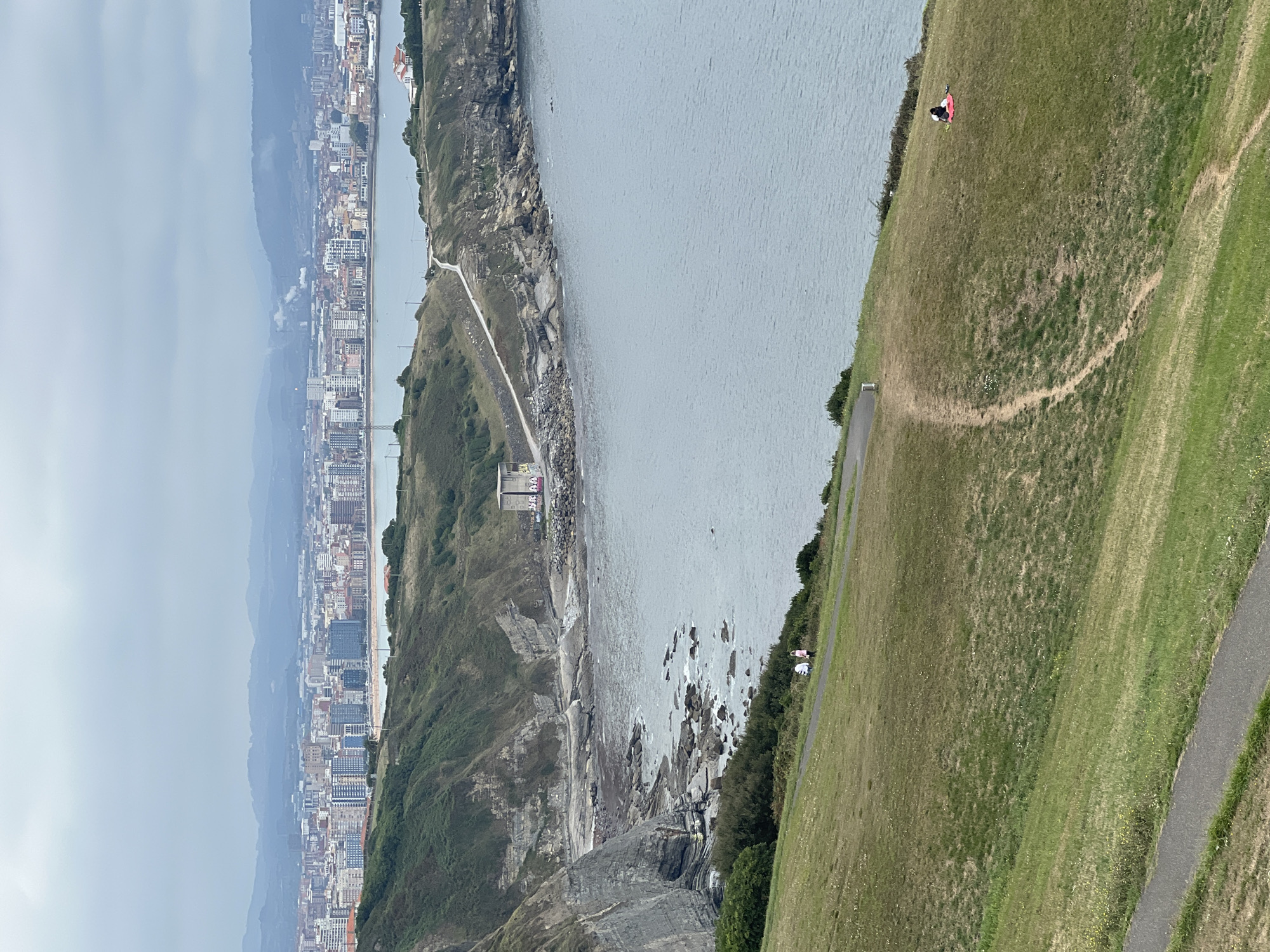 The image captures a panoramic view of the coastline in Gijón, Spain, with the city skyline in the distance. The foreground features a lush green hillside with a paved path winding through it. Two individuals are visible walking on the path on the left side of the image, and a solitary person is seated on a red blanket on the grassy slope on the right. The middle ground showcases a rugged, rocky coastline meeting the calm blue sea. A narrow road traverses the cliffside, leading to a modern, square building with graffiti. In the background, the urban landscape of Gijón unfolds, characterized by a dense arrangement of multi-story buildings stretching along the coast, with a hazy, mountainous horizon beyond. The sky is overcast, contributing to a subdued, natural light.