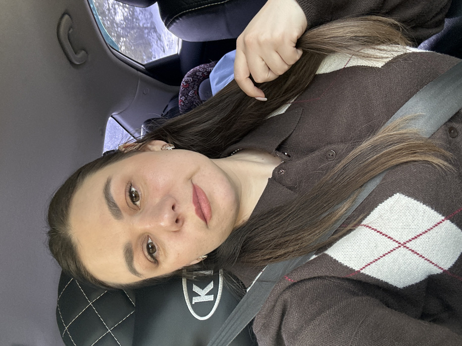 This image depicts a female subject seated inside a vehicle. The scene is identified as being in Krasnye Gorki, Russia. The subject, a woman with long, dark brown hair parted to her left, is looking directly forward towards the camera. She has light-colored eyes, defined eyebrows, and appears to be wearing subtle makeup, including a light pink lip color. A small, ornate earring is visible in her right ear. She wears a brown collared cardigan featuring a white and maroon argyle pattern on the front, with buttons running down the center. A silver-toned chain necklace is visible around her neck. A gray vehicle seatbelt diagonally crosses her chest. Her right hand is shown lightly grasping a section of her hair near her shoulder.
The immediate environment is the interior of a car. A black, quilted headrest with a white "KIA" logo is partially visible behind her left shoulder. In the background, beyond the subject, the vehicle's rear interior shows parts of a dark seat, a blue object, and a patterned child car seat. Through the rear and side windows, a distinct snowy landscape is visible, characterized by snow-covered ground and dark, bare trees, indicative of a winter outdoor environment. The car's ceiling and a grab handle are also partially visible.