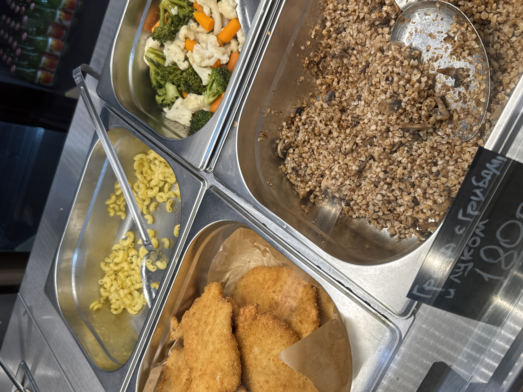 The image shows a food display case in a cafeteria. There are four metal containers filled with different food items. The top left container has cooked pasta. The top right container has steamed vegetables, including broccoli, cauliflower, and carrots. The bottom left container holds several breaded and fried cutlets. The bottom right container contains buckwheat with mushrooms. A metal scoop is visible in the buckwheat. A price tag is partially visible in the lower left corner, with handwritten Cyrillic text that appears to read "Гречка с грибами" (Buckwheat with mushrooms) and "180" followed by what looks like rubles. The environment appears to be a food service establishment. There are no people clearly visible in the image. The lighting is bright, suggesting an indoor setting. The overall impression is that of a self-service food line.
