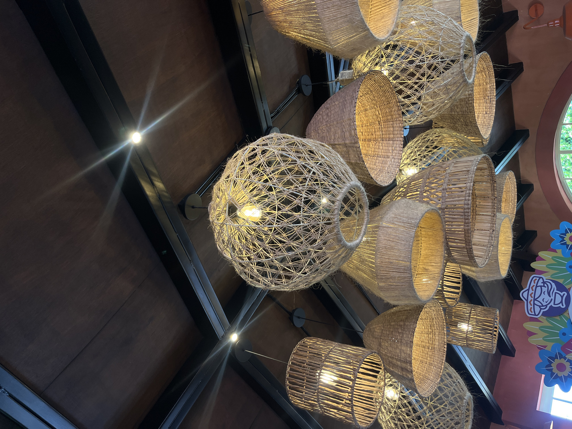 The image shows a view looking up at a ceiling adorned with numerous woven basket-style light fixtures. The ceiling itself is constructed of dark wood panels intersected by black metal beams, with recessed lights positioned along the beams. The light fixtures vary in shape and size, including round and cylindrical forms, all crafted from a natural fiber material. The fixtures are illuminated, casting a warm light. In the background, hints of a red-orange wall and a window are visible, alongside a decorative sign with Mexican-themed graphics. The location is suggested to be in Puebla, Mexico.