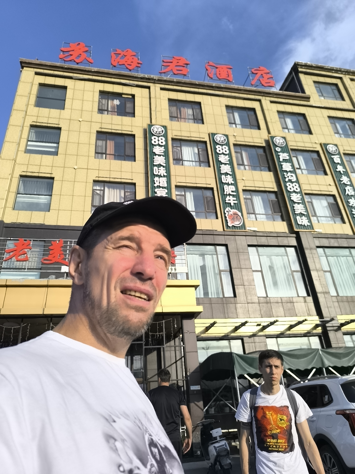 In a low-angle shot taken outdoors in front of a multi-story building, a man wearing a white t-shirt and a black baseball cap is in the foreground, looking to the right and smiling. Behind him, a younger man wearing a white t-shirt with a graphic print and a backpack stands facing the camera. Two white SUVs are parked behind the younger man, and a motorcycle is to his left. The building in the background is yellow with red Chinese characters at the top that read "春海酒家," which translates to "Chunhai Restaurant." Vertical green banners with Chinese text are also visible on the building's facade. The sky is blue with some clouds. The scene appears to be in Lucaogou Town, China.