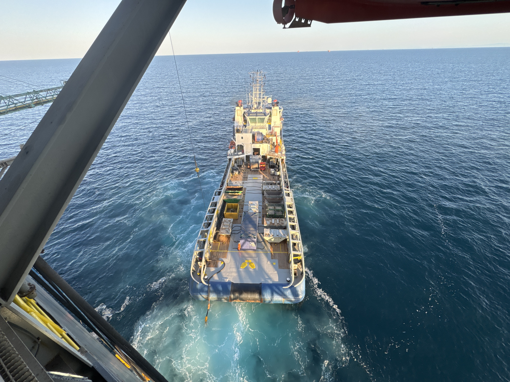 A high-angle, medium shot captures a view from a vessel looking down at another vessel moving through calm, deep blue ocean waters under a clear, pale sky. The secondary vessel, a supply ship, is positioned in the center of the frame, its stern facing the viewer. The ship has a wide, flat deck covered with various containers and equipment, including several open-topped bins and what appear to be cargo nets. A bright yellow "X" is visible on the deck towards the stern. A hose extends from the supply ship into the water, creating a white foamy wake as the vessel moves. The surrounding ocean is a rich, deep blue with small ripples on its surface, transitioning to a brighter turquoise near the ship's wake. The foreground is partially obscured by the dark grey, angled structure of the viewing vessel's railing and a red-painted overhead element. The horizon line is distant and clear.