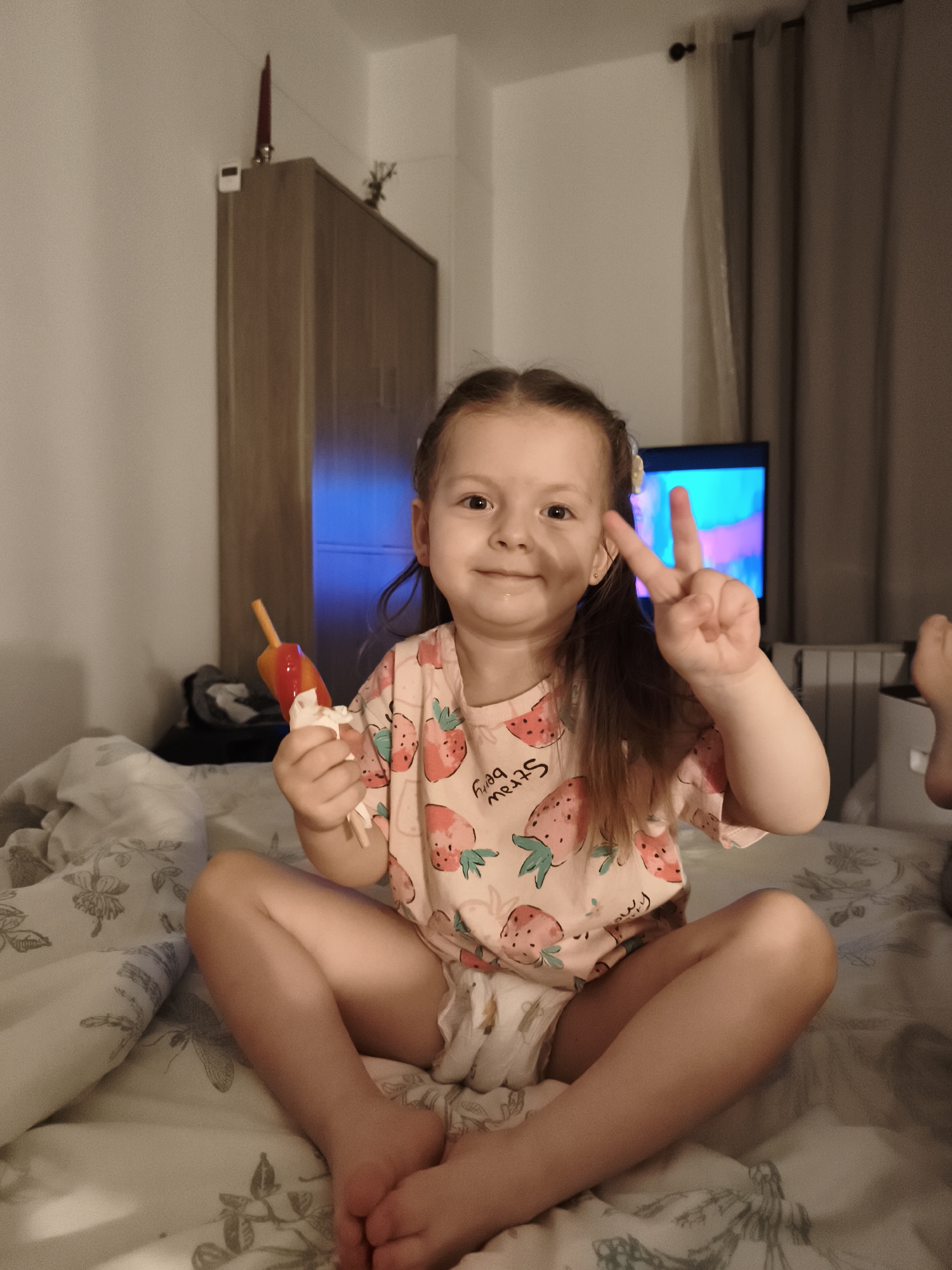 A young girl with brown hair is sitting cross-legged on a bed. She is wearing a pink t-shirt with a strawberry pattern and a diaper. In her right hand, she holds a popsicle. She is smiling and making a peace sign with her left hand. The background shows a wardrobe, a television screen with colorful content, and curtains. The scene appears to be indoors, likely a bedroom, during the evening or nighttime, as indicated by the artificial lighting and the dim ambiance. The girl's expression conveys happiness and playfulness. The t-shirt has visible text that reads "STRAWBERRY."