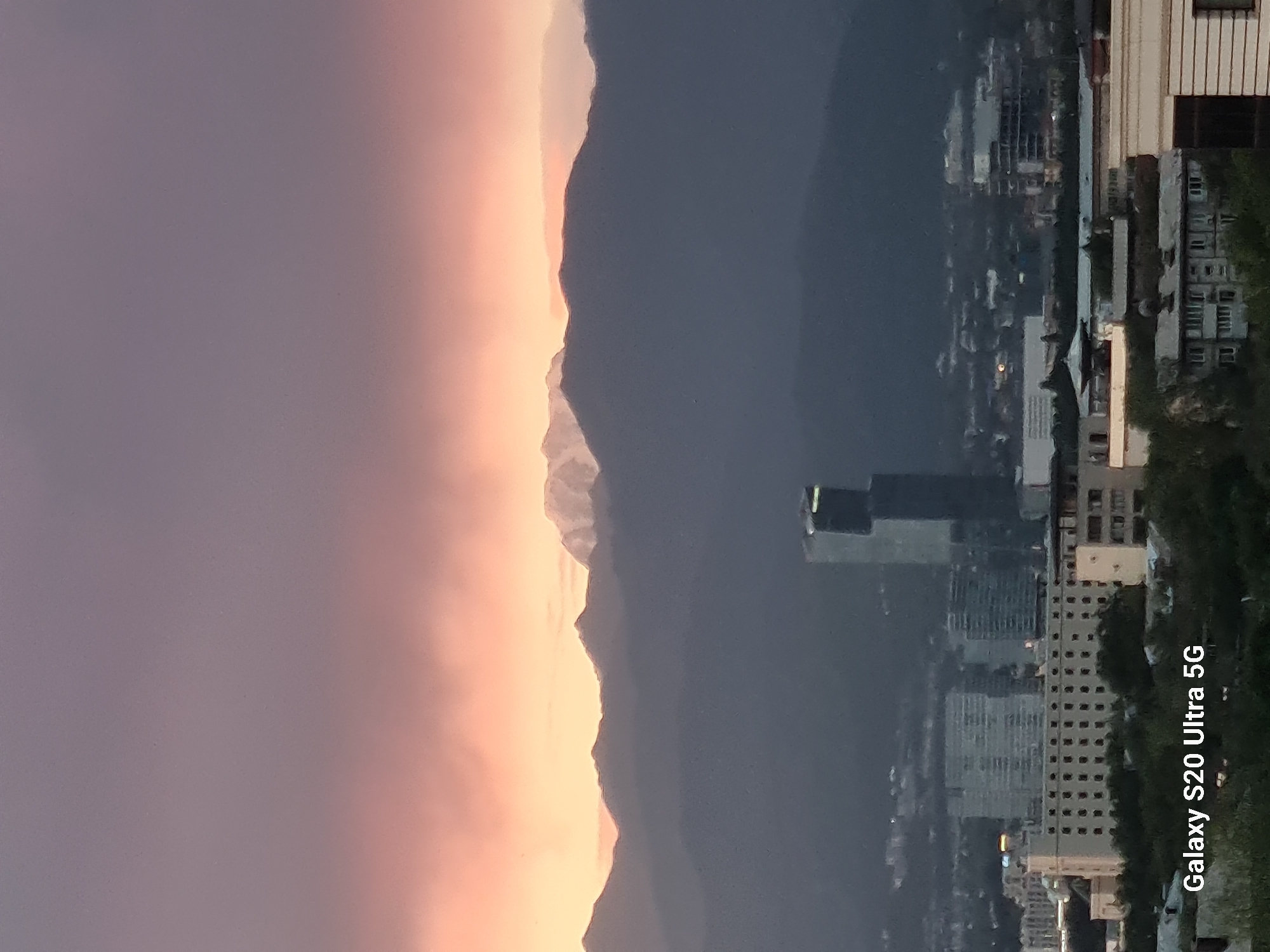 The image captures a panoramic view of Almaty, Kazakhstan, at dusk. The sky is painted with the soft hues of sunset, transitioning from a peachy orange at the horizon to a muted purple and gray above. In the background, majestic, dark mountains loom, their peaks dusted with snow and illuminated by the fading light.
Nestled at the foot of the mountains, the city of Almaty unfolds. Modern high-rise buildings, including a striking glass skyscraper with a distinctive angular design, punctuate the urban landscape. Lower-rise buildings, apartment complexes, and what appear to be administrative or commercial structures are visible, their windows reflecting the ambient light. Greenery, likely parks or tree-lined streets, can be seen interspersed among the buildings, adding a touch of nature to the concrete jungle. The foreground is dominated by trees and rooftops, suggesting a vantage point from a higher elevation within the city. A watermark at the bottom left corner of the image reads "Galaxy S20 Ultra 5G," indicating the device used to capture the scene. The overall impression is one of a serene urban environment embraced by the grandeur of nature.