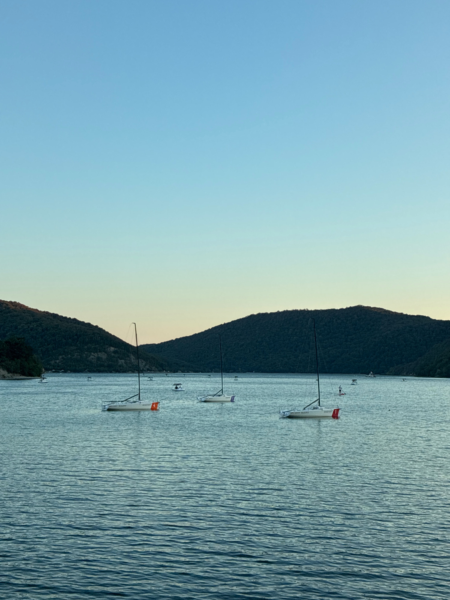 The image depicts a tranquil scene on a body of water, likely a lake or a bay, with rolling, tree-covered hills in the background. Several sailboats are visible on the water, some with sails down and others with masts raised. One sailboat in the foreground has the number "4" and the word "RUSSPU" visible on its hull. The sky is a clear, pale blue, transitioning to a lighter hue near the horizon, suggesting either early morning or late afternoon. The water is calm, with subtle ripples reflecting the sky and surrounding landscape. In the distance, more sailboats and what appear to be small buoys or markers are scattered across the water. The overall atmosphere is one of peacefulness and calm, characteristic of a natural recreational area. This scene is consistent with views of the Abrau Liman, a freshwater lake near the village of Abrau-Dyurso in Krasnodar Krai, Russia, which is known for its sailing activities and picturesque natural surroundings.