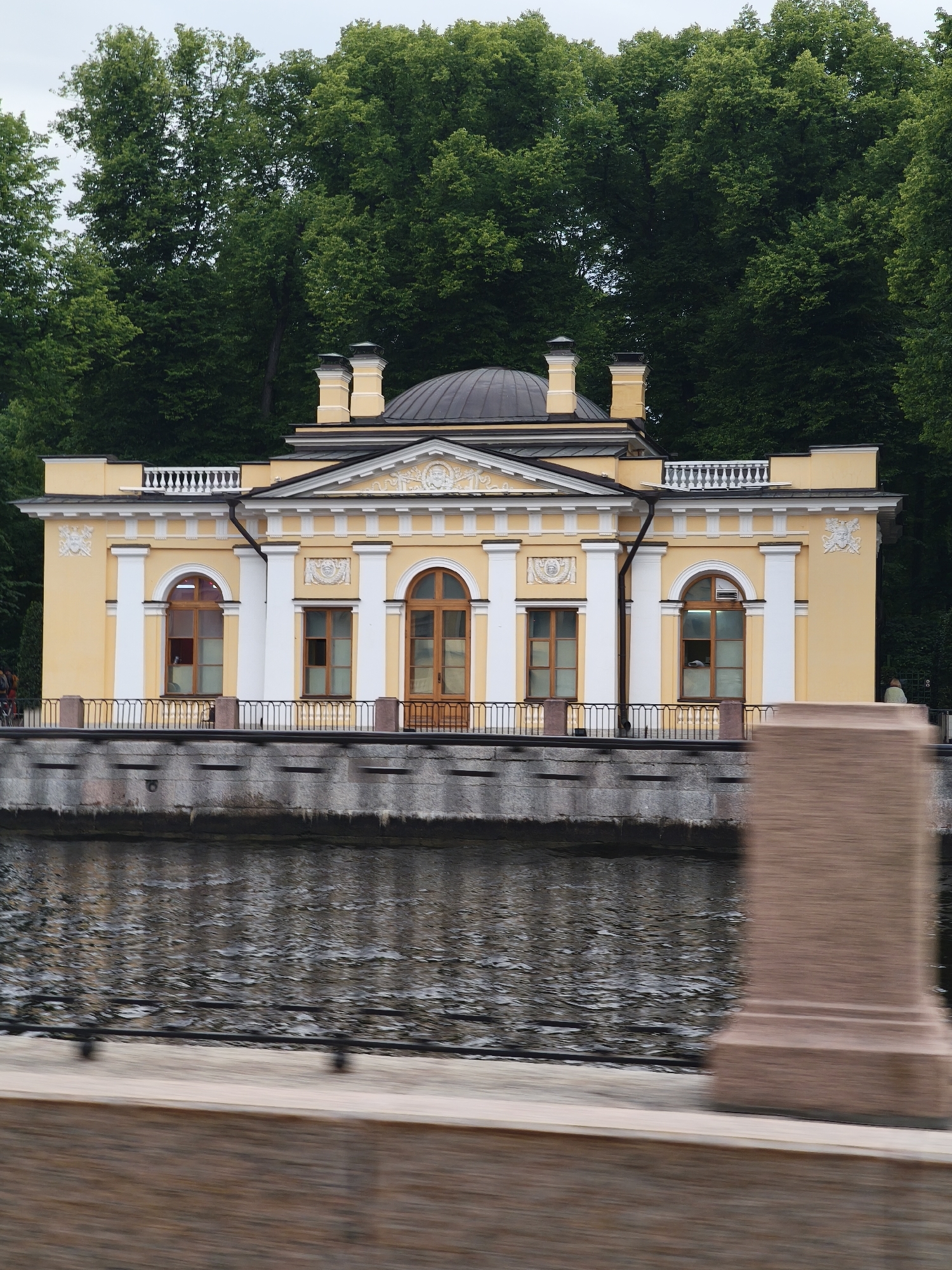 The image depicts a yellow neoclassical building with white columns and decorative elements, situated along a stone embankment of a dark, rippling body of water. The building features a central dome and arched windows with wooden frames. Lush green trees form a dense backdrop behind the structure, suggesting a park or garden setting. A black metal railing runs along the top of the embankment, separating the building from the water. In the foreground, a blurred, reddish-brown structure, possibly a monument or part of a bridge, is visible, creating a sense of movement or depth.
The scene appears to be set in daylight, with overcast or slightly diffused lighting, indicated by the soft shadows and the sky's appearance. There are hints of people near the building on the left, though they are small and indistinct, and a single person is visible on the far right of the embankment. The overall atmosphere is serene and picturesque, conveying a sense of historical grandeur. The location context of Saint Petersburg, Russia, aligns with the architectural style, which is typical of many historical buildings in the city. No visible text is present in the image.