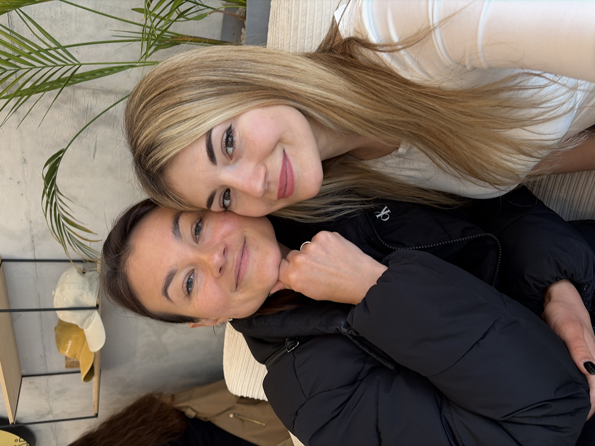Two smiling women pose closely together indoors, likely in a cafe or restaurant setting. The woman at the top has long blonde hair and is wearing a light-colored shirt. The woman below has brown hair and is wearing a black jacket, which features the Calvin Klein logo on the chest. Her left hand is placed under her chin, while her right hand rests on her lap. A palm tree branch is visible in the upper left corner. To the left of the women, there's a shelf with several hats on it. The photograph seems to have been taken indoors in a place in the city of Budva, Montenegro.