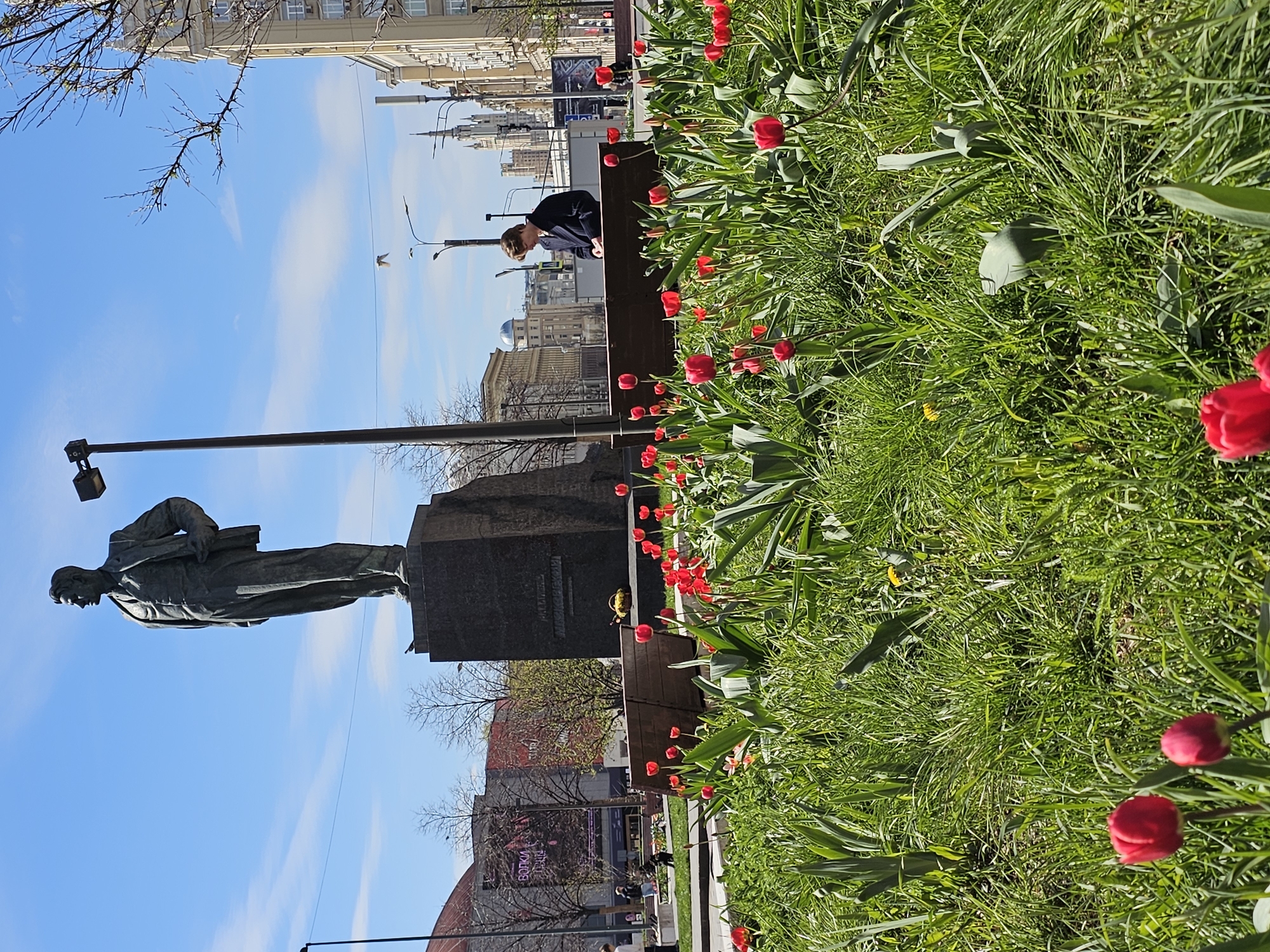 The image shows a statue, most likely a monument, in what appears to be a park-like setting with tulips blooming in the foreground. The statue is of a man in a suit with a coat draped over his arm and a book in his hand, standing on a pedestal. In the background, there are buildings typical of a city landscape. A person can be seen sitting on a bench near the monument. The sky is blue with scattered clouds. The location is Moscow, Russia.