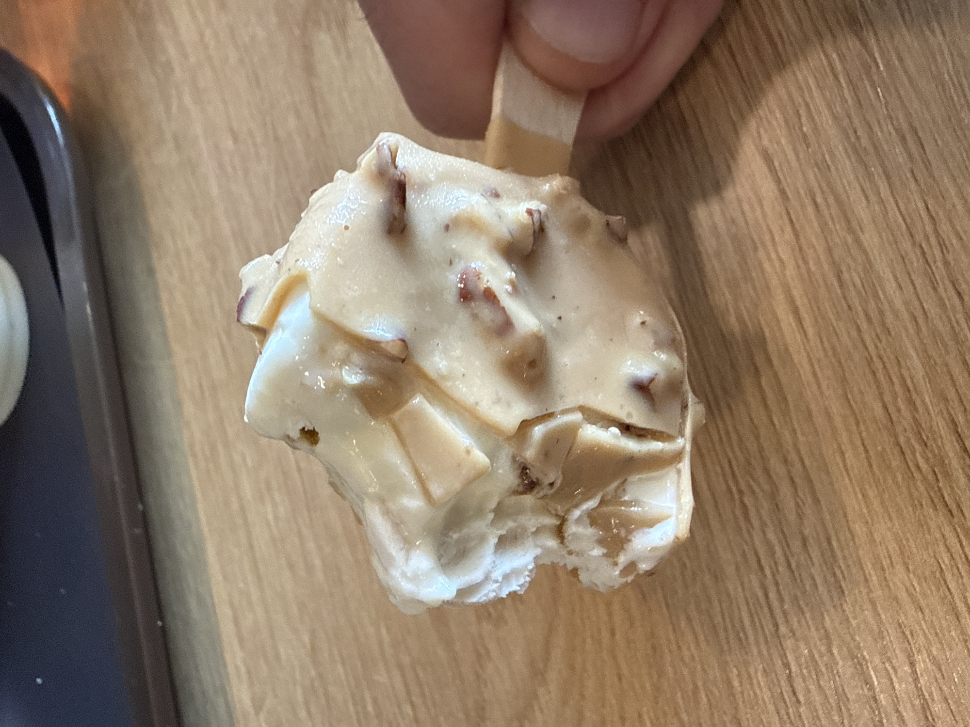 A partially eaten ice cream bar on a wooden stick is held by a person's thumb and finger. The ice cream has a light brown coating with embedded pieces, possibly nuts. A bite has been taken from the bottom of the ice cream bar revealing a white interior. A dark-colored tray is visible on the left. The surface underneath the ice cream bar is a light brown woodgrain. No information in the image indicates the location is Sofia, Bulgaria.