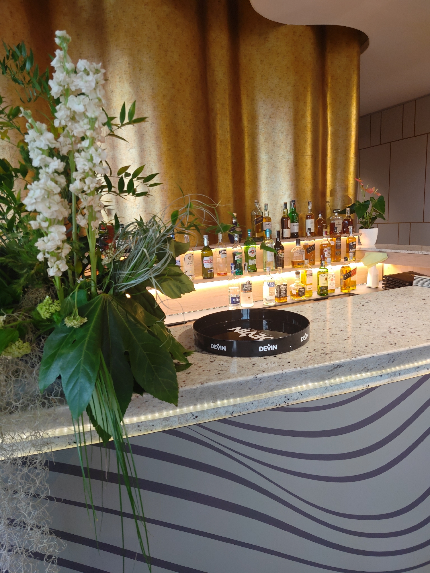 The image shows a bar in a possibly upscale setting. A large floral arrangement dominates the left side of the frame. Behind it, liquor bottles are arranged on a tiered counter with visible under-lighting. A black tray with "DEVIN" branding sits on the countertop. The counter has a speckled surface and a wavy, grey and white pattern on its front. A golden textured wall rises behind the bar area. The setting appears to be indoors, and there is a small plant in a white pot on the top shelf towards the right. There is no immediately evident indication of the city of Varna, Bulgaria from the imagery.