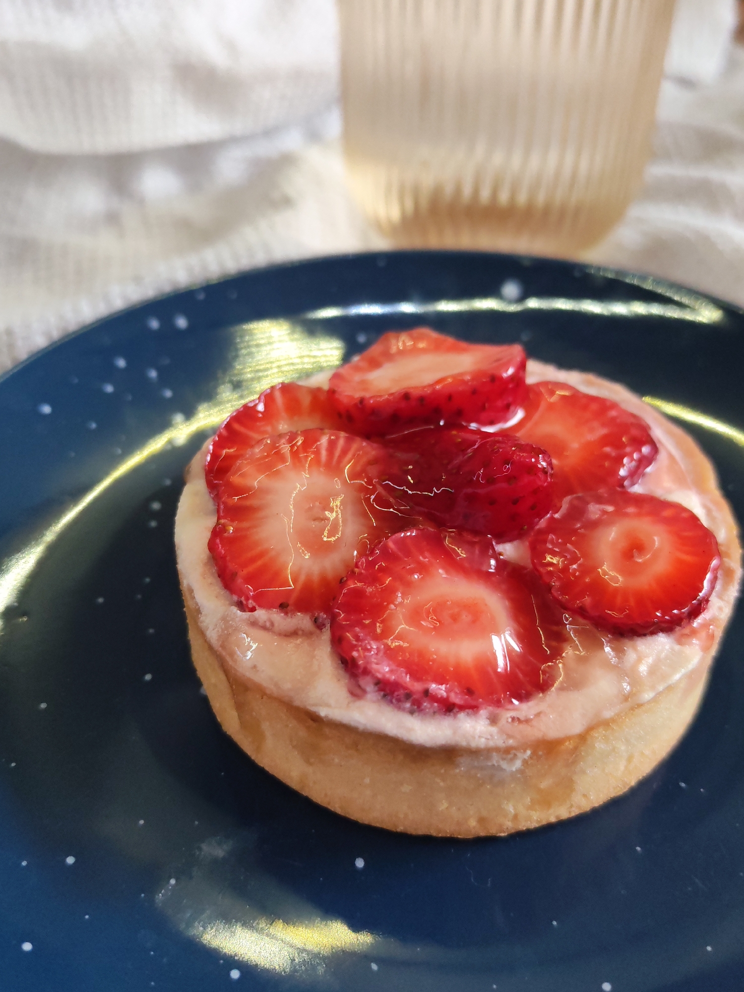 The image shows a strawberry tart on a dark blue plate with white speckles. The tart consists of a shortbread crust, a layer of light-colored cream, and sliced strawberries glazed with a clear, shiny coating. A glass with a light amber liquid is partially visible in the background. A white fabric with wrinkles lies behind the plate. The location is not discernible from the image.