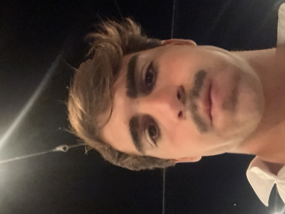 A low-angle selfie captures a fair-skinned person with brown hair styled with volume. The individual sports exaggerated, drawn-on eyebrows and mustache. The background is dark, suggesting nighttime. A blurred, bright light appears on the left. The person wears a white garment. The overall composition is intimate and close-up.