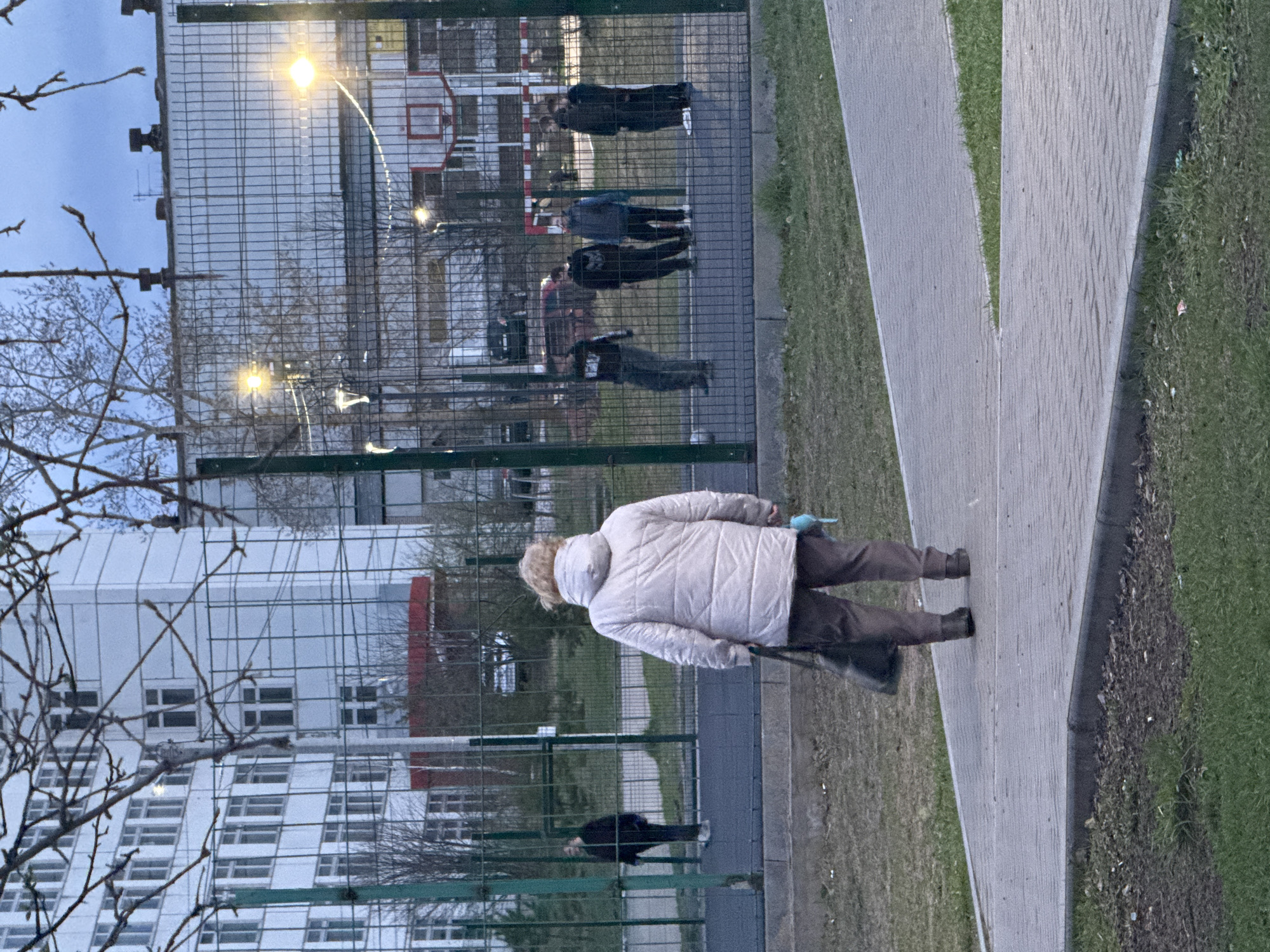 The image, taken in Chaikovsky, Russia, captures an outdoor scene at what appears to be dusk or dawn, given the subdued light and illuminated streetlights. The cool weather is suggested by bare tree branches in the foreground and warm clothing worn by the subjects.
In the immediate foreground, an older individual with light-colored hair, wearing a cream or white puffer jacket and dark trousers, walks away from the viewer on a grey, textured paved path. They are holding what appears to be a dark bag or walking stick, and a light blue object (possibly a glove or mask) is visible near their jacket collar.
A tall, dark mesh fence with horizontal green beams runs parallel to the path, separating it from an inner area. Behind this fence, several other people are visible. Approximately four younger individuals are standing on what looks like a sports court, distinguished by a visible red basketball hoop and striped markers. Their activity is casual, suggesting they are gathered or lingering. Further in the background, another person in dark clothing is visible near a building.
The background consists of large, multi-story residential buildings with numerous windows, reflecting an urban or suburban setting. One building has a distinctive white and grey facade with angled sections, while another features a prominent vertical red strip. Two glowing streetlights behind the fence provide additional illumination. Green grass borders the paved pathway and is also present within the fenced area. No visible text is discernible in the image.