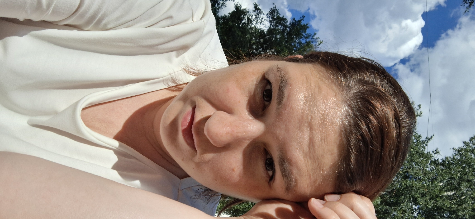 A woman with reddish-brown hair and fair skin is looking directly at the camera, with her right hand touching her forehead. Her head is tilted slightly downwards. She is wearing a white, scoop-neck t-shirt. The background consists of a bright blue sky with fluffy white clouds and a blurred green tree. The lighting suggests it is daytime and sunny. There are some freckles visible on her face. The image appears to have been taken outdoors in a location that could be Chaikovsky, Russia, given the presence of deciduous trees common to the region.