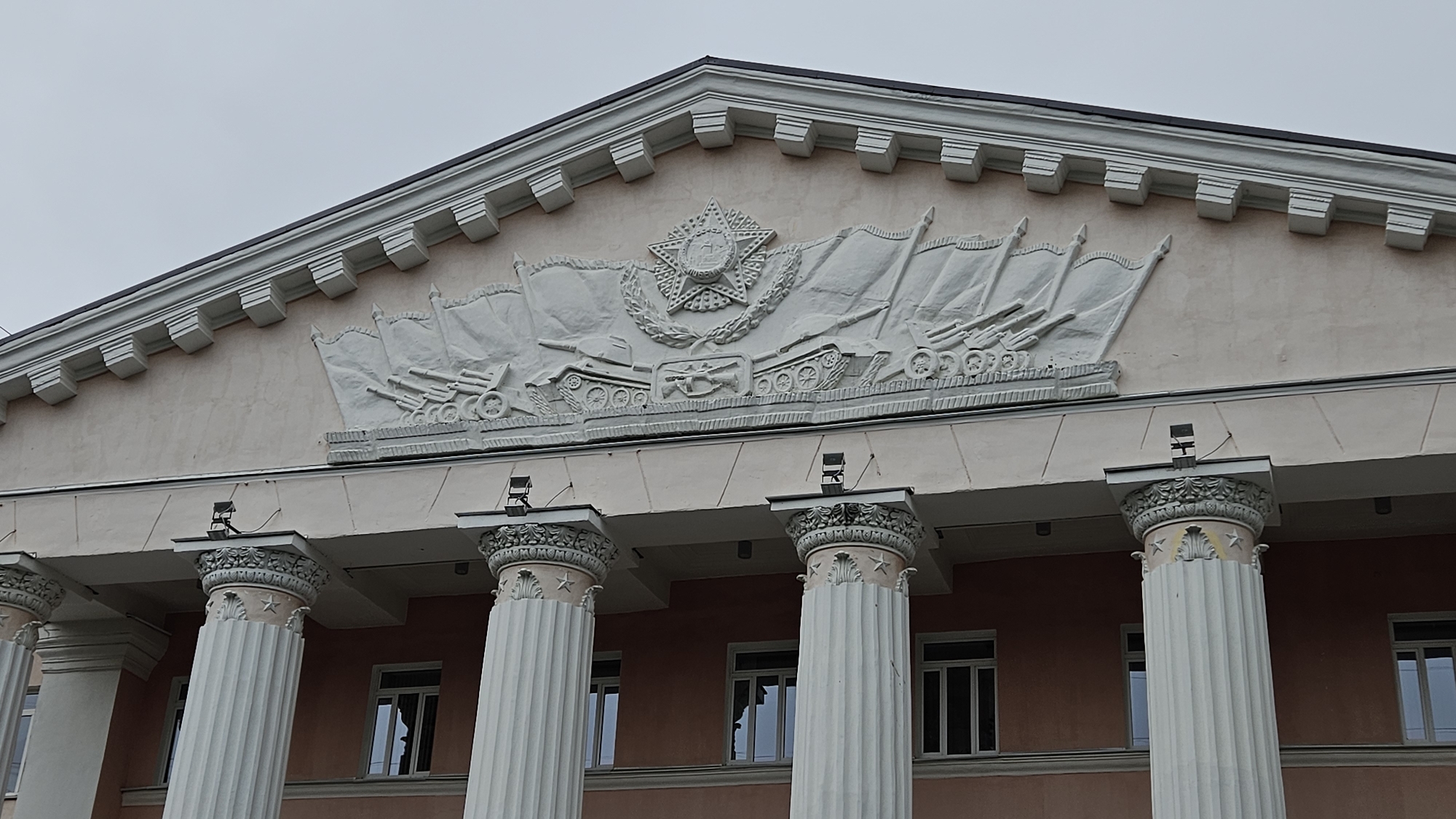 The image shows a building with neoclassical architectural elements. The facade is a light pink color and features four fluted columns with ornate capitals. Above the columns, there is a frieze topped with a large relief sculpture featuring military motifs, including a five-pointed star surrounded by a wreath, tanks, artillery guns, and flags. The sky is overcast and grey. Windows are visible behind the columns. The city and country are not discernible from the image.