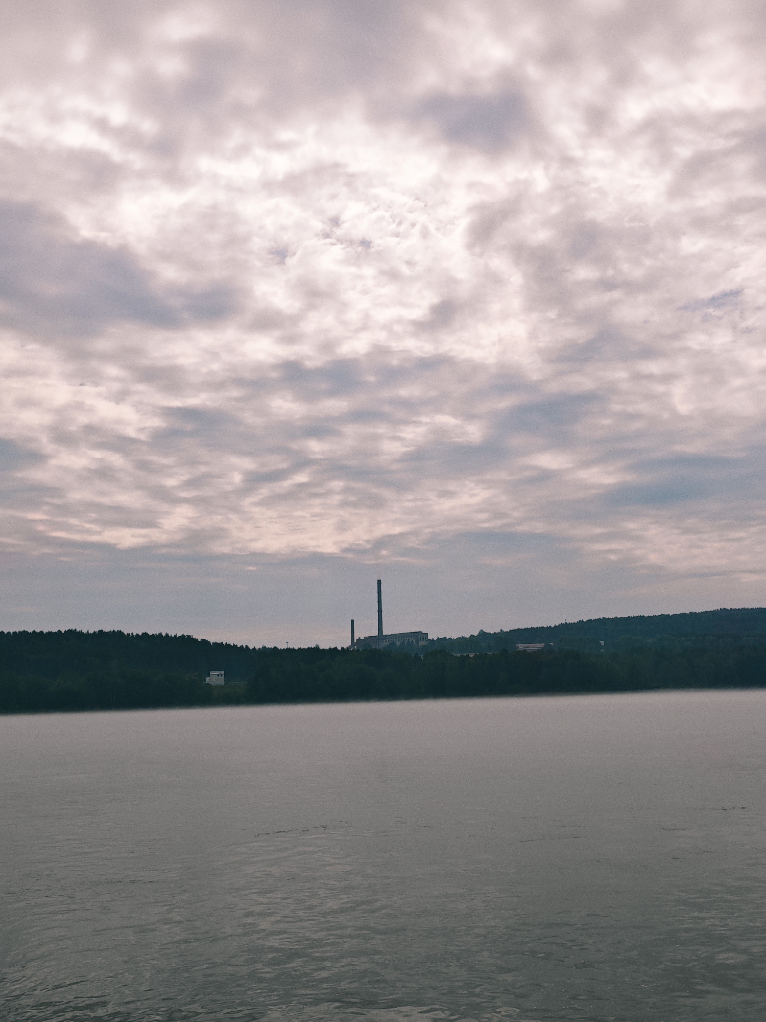 The image depicts a wide body of water, likely a river, in Atamanovo, Russia, under a cloudy sky. The water's surface is calm, with subtle ripples and a slight haze or fog near the distant shore. The shoreline is densely forested with dark green trees. On the elevated bank, there is a cluster of industrial buildings with tall smokestacks, suggesting a power plant or factory. To the left of the industrial complex, a small, white, rectangular building stands out against the dark foliage. The sky is filled with a thick layer of grey and pinkish clouds, creating a muted and somewhat overcast atmosphere. The overall scene suggests an industrial area situated by a natural watercourse.