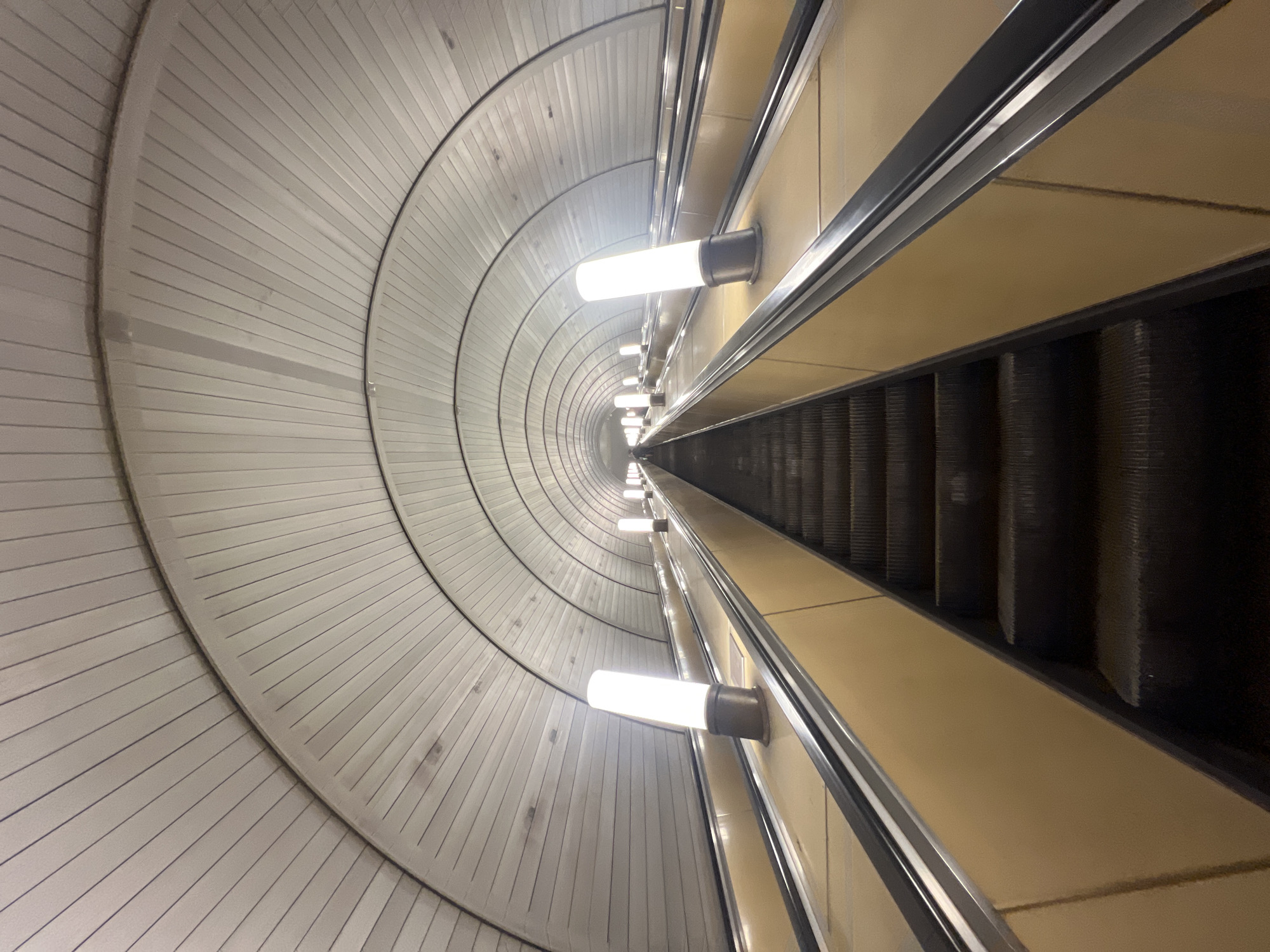 The image depicts an escalator descending into the Moscow Metro in Russia. The architectural style is characterized by a large, rounded ceiling structure with repeating radial panels that narrow into the distance. This ceiling is illuminated by a row of cylindrical light fixtures mounted along the side. The escalator itself is made of dark metal steps, flanked by beige-colored walls and metallic handrails. The scene is viewed from a high angle looking down the length of the escalator. No people are visible in the image.