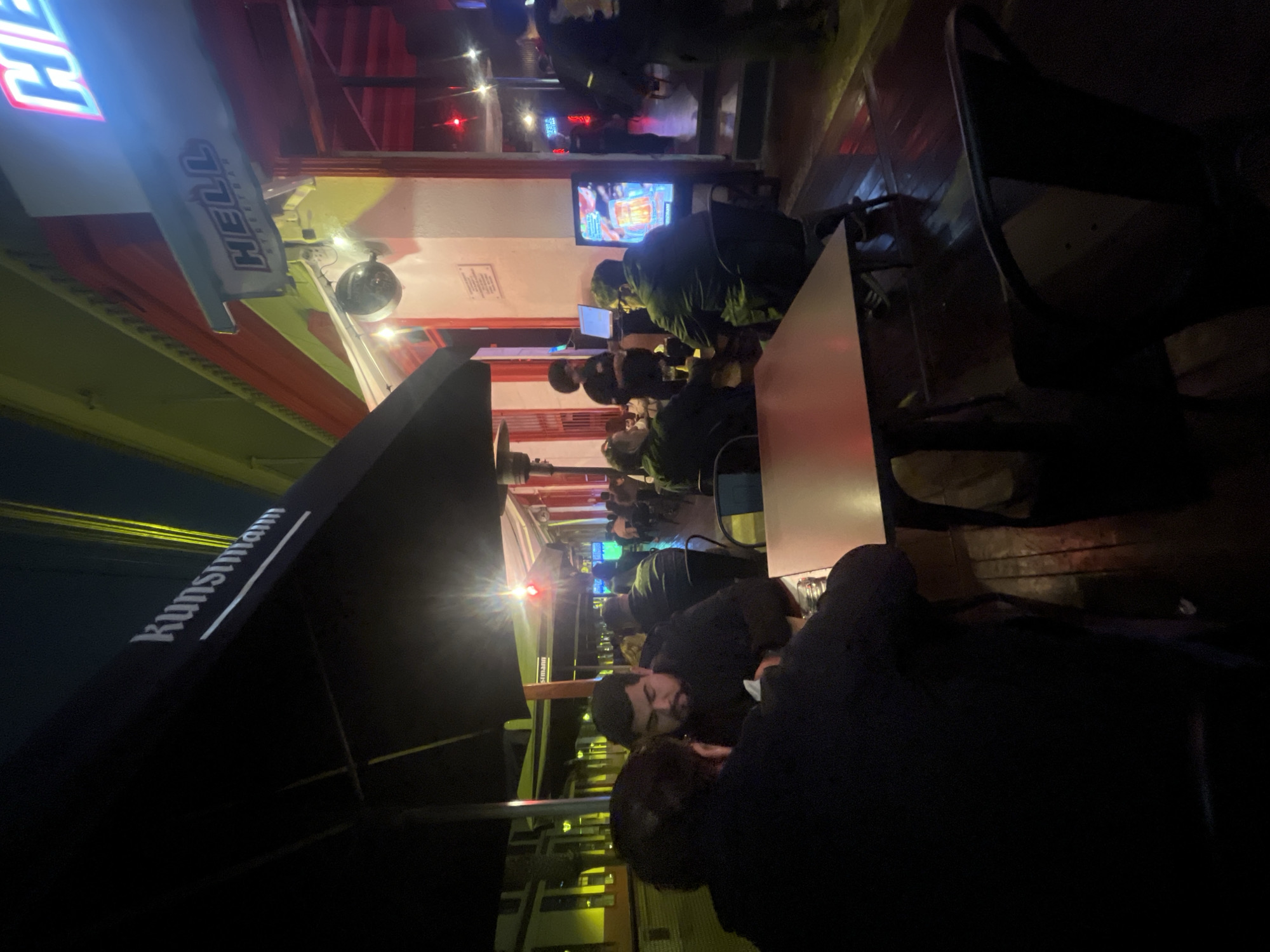 The image depicts an outdoor seating area of a restaurant or bar at night in Iquique, Chile. The establishment features a prominent sign reading "HELL STREETBAR" illuminated with neon lights. Another sign, partially visible and partially obscured by a black awning, reads "Kunstmann" with a white underline. Several patrons are seated at tables, some engaged in conversation, while others appear to be observing the surroundings. One individual is standing near a television screen displaying colorful graphics. The lighting suggests an evening scene, with artificial lights from the establishment and streetlights casting a warm glow. The ground appears to be paved, with some wet patches suggesting recent rain. The overall atmosphere is that of a lively urban nightlife scene.