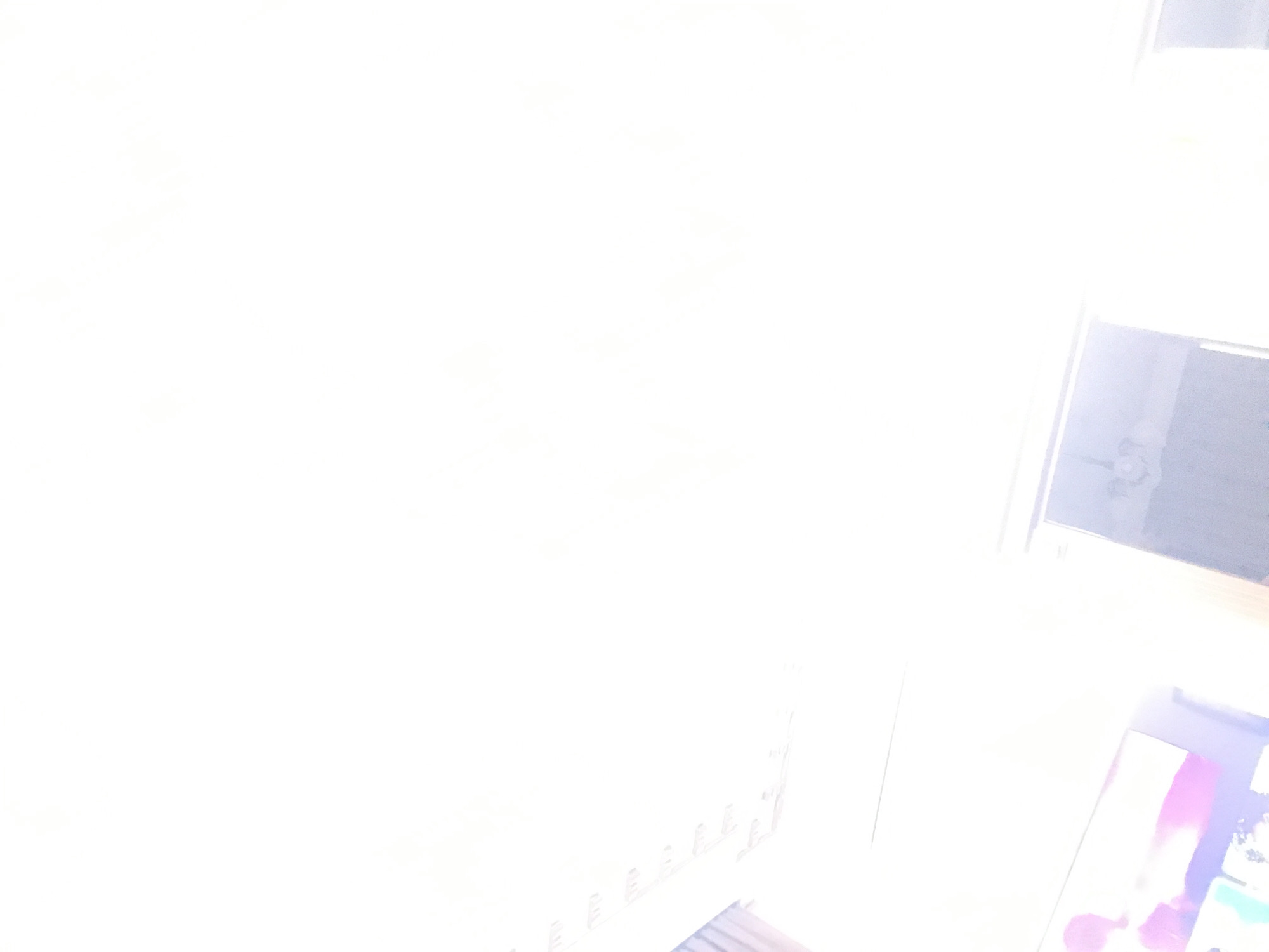 The image is severely overexposed, making it difficult to discern details. However, discernible elements include: part of a TV screen with purplish tones in the lower right corner, a window with a blurry view outside on the upper right of the image, what appears to be the edge of a shelf, a blurry set of books along the lower part of the image. There is not enough information to determine the location in Yerevan, Armenia.