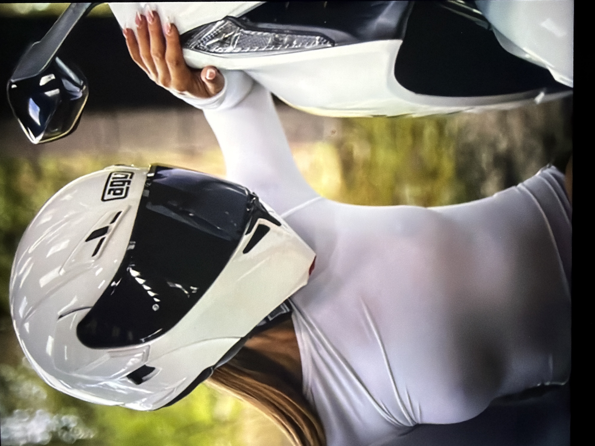 The image displays a close-up, portrait-oriented shot of a person posing outdoors with a motorcycle and a helmet. The person, whose lower face is obscured by the helmet and upper body visible, is wearing a snug, long-sleeved white top. Long, light-colored hair (blonde or light brown) is visible flowing from beneath the helmet. A hand with manicured, light pink nails is gently placed on a white section of a motorcycle, with a black rearview mirror also visible to the left.
A prominent white AGV brand motorcycle helmet, featuring a dark visor and black ventilation accents, is positioned on or near the person's head. The brand name "AGV" is clearly legible in black lettering on a white emblem on the side of the helmet.
The background is softly blurred, presenting tones of green and yellow, indicating a natural, outdoor setting with foliage or trees. The natural lighting suggests it is daytime. The scene conveys a stylish or promotional aesthetic. No specific location cues referencing Hamburg, Germany, are visible within the image itself.