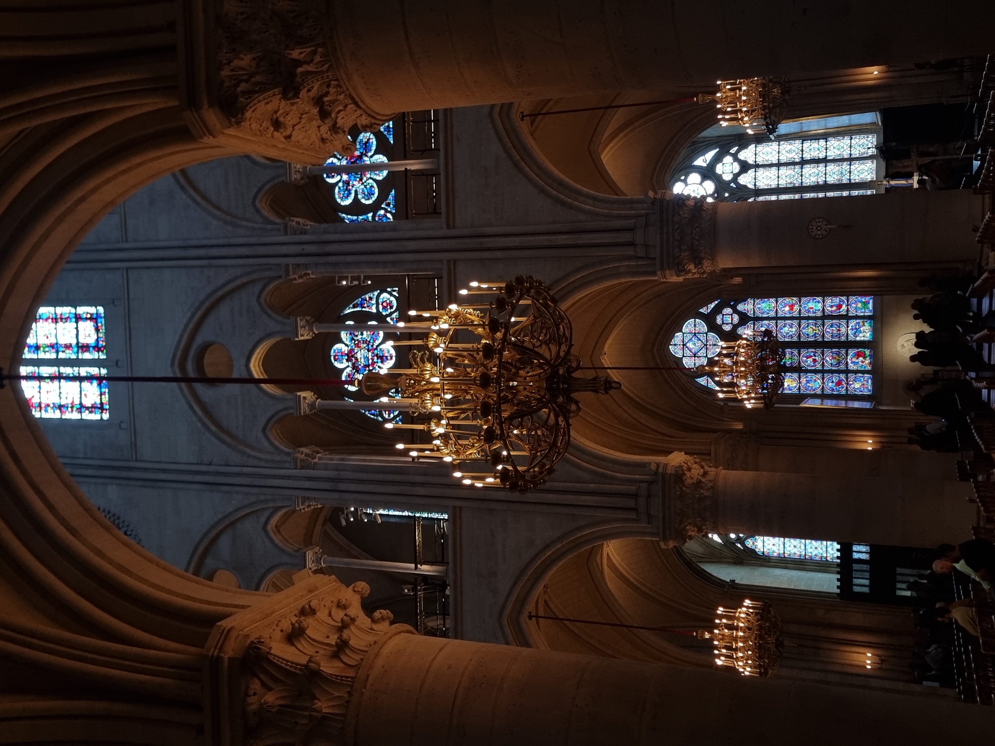 This image captures the majestic interior of a grand cathedral or church, characterized by its towering Gothic architecture. The perspective, which appears to be rotated 90 degrees clockwise, looks across an aisle towards the side walls and ceiling.
Dominating the scene are massive stone columns and intricate ribbed vaulted ceilings, forming elegant arches overhead. Several large, ornate chandeliers, crafted from golden-hued metal and fitted with numerous electric candles, hang from the ceiling, casting a warm, luminous glow that illuminates the vast space.
Multiple stained glass windows are visible along the upper walls. These include large circular rose windows and rectangular panels, showcasing vibrant geometric and floral designs in blues, reds, and other rich colors. These windows allow natural light to filter into the otherwise dimly lit interior, creating a solemn and awe-inspiring atmosphere.
On the right side of the image, a group of people is visible, mostly in silhouette or softly illuminated. They appear to be seated or standing in dark wooden pews or benches, observing their surroundings. Their postures suggest a reverent or contemplative mood, indicative of attending a service or touring a historic site.
The architectural style, featuring soaring arches, vaulted ceilings, and elaborate stained glass, points to a significant European cathedral. Given the context of Paris, France, it strongly resembles a major landmark like Notre Dame Cathedral. The lighting suggests either a heavily overcast day or an interior that relies significantly on artificial illumination, creating a dramatic and serene ambiance. No visible text is present.