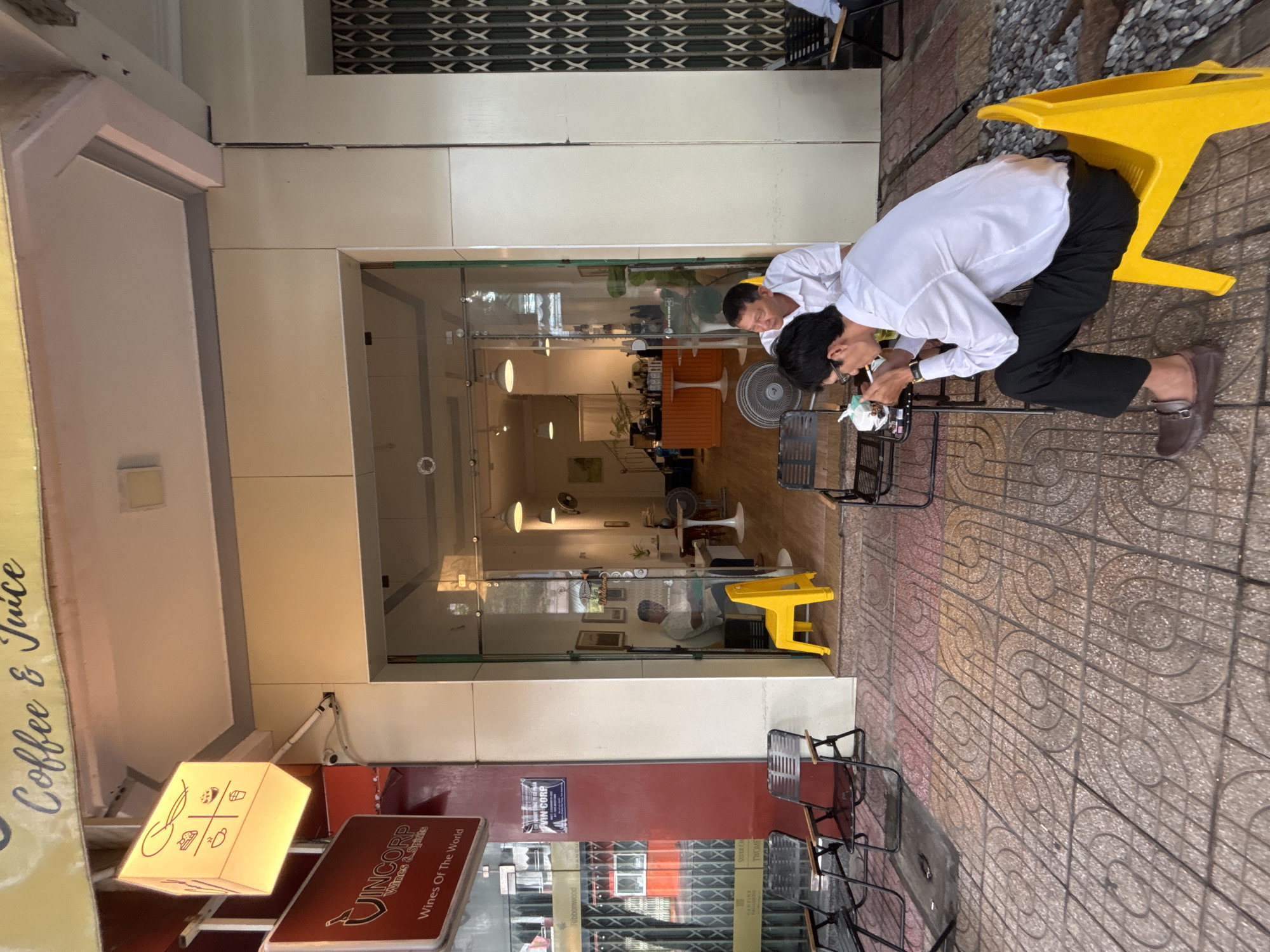 The image shows a street-level view of a coffee shop in Nha Trang, Vietnam. The coffee shop has a bright yellow illuminated sign with icons of food and drink. Below it is a red sign for "VINCORP Wines Of The World". The entrance to the shop is open, revealing a well-lit interior with tables, chairs, and a counter.
In the foreground, two men are seated at a table on the sidewalk. One man, wearing a white shirt and black pants, is smoking a cigarette and looking down at his drink. He is sitting on a bright yellow plastic chair. The other man, also in a white shirt, is seated behind him and looking to the side. There are other outdoor seating options visible, including black metal chairs and tables. The ground is paved with patterned tiles. The overall scene suggests a casual outdoor seating area for a coffee shop.